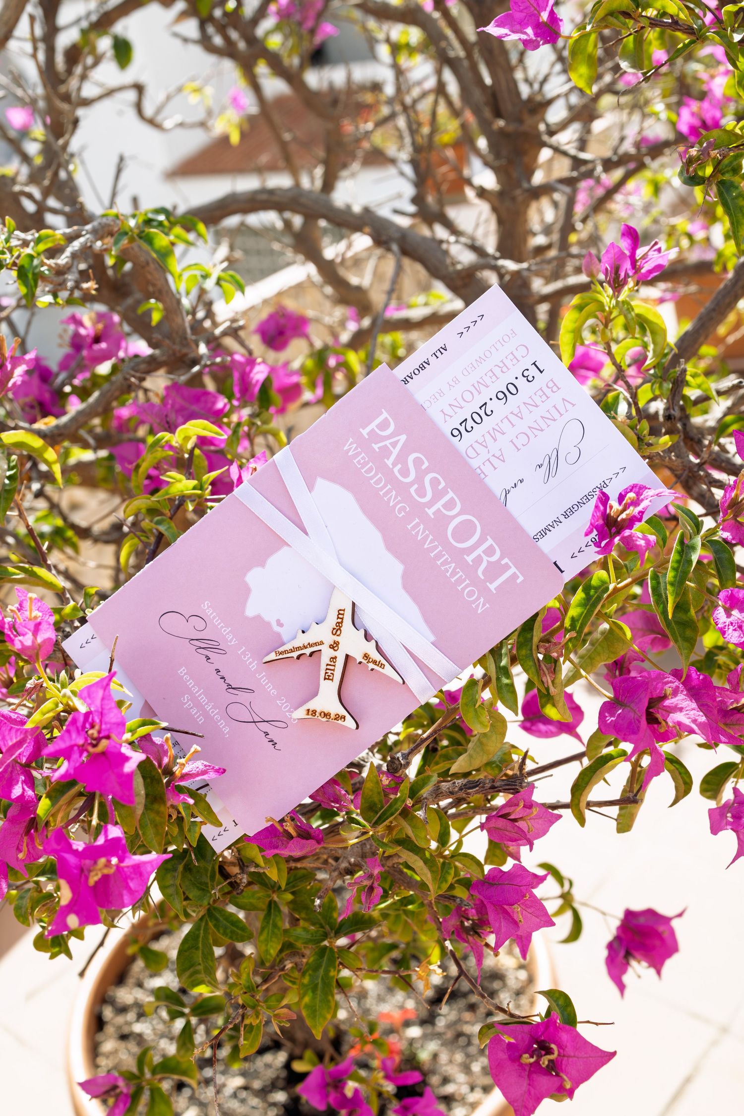 Pink passport wedding invitation with airplane charm displayed among vibrant bougainvillea flowers in bright sunlight.