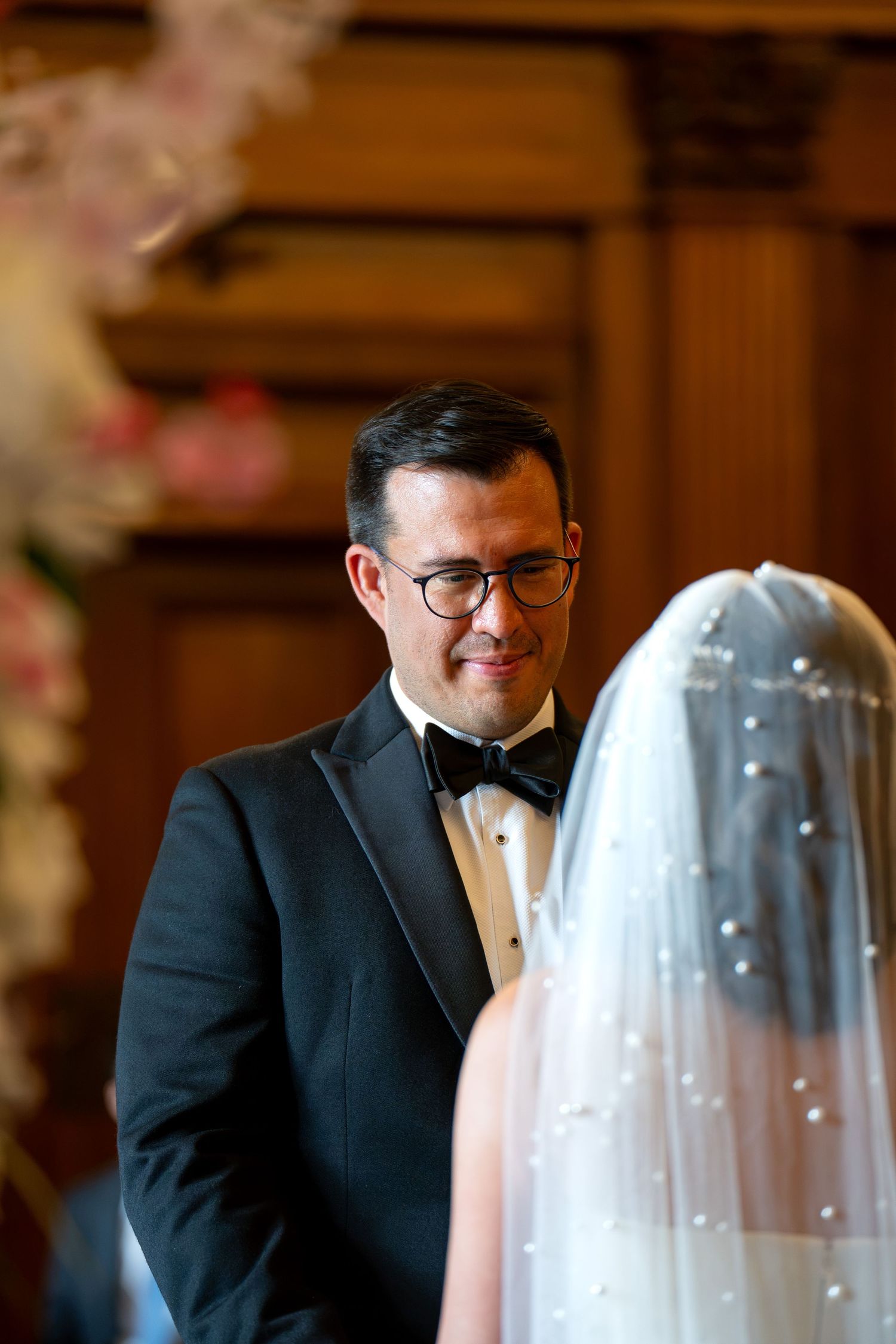 Emotional groom in black tuxedo and glasses watches his bride during wedding ceremony indoors.