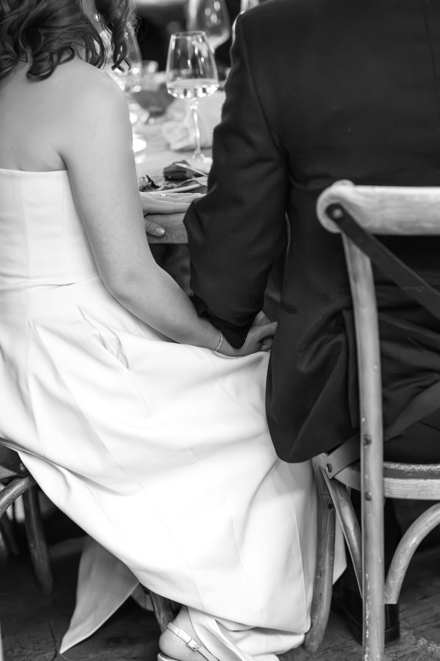 Black and white photo of bride and groom holding hands intimately while seated together at wedding reception table.