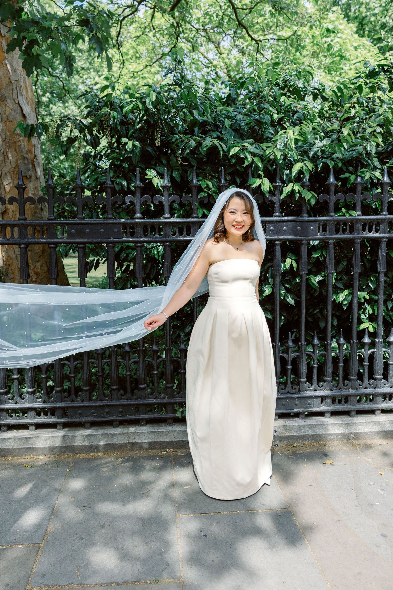 A bride in a strapless white gown lets her veil flow in the breeze beside an ornate black iron fence.