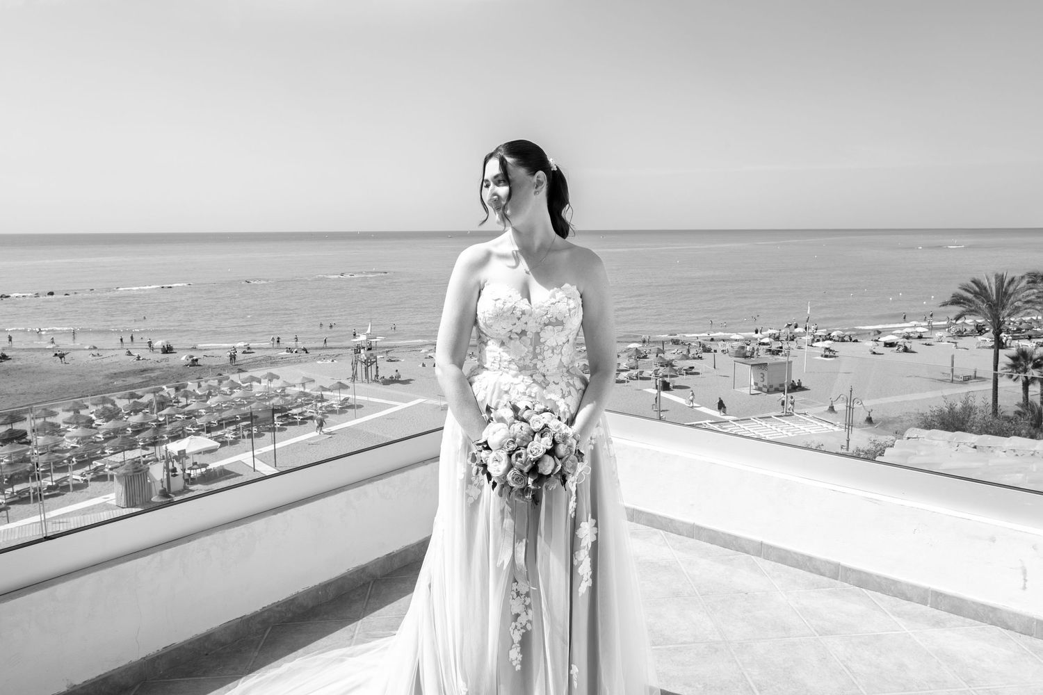 Bride in strapless gown holding bouquet on seaside terrace with beach and palm trees in black and white.
