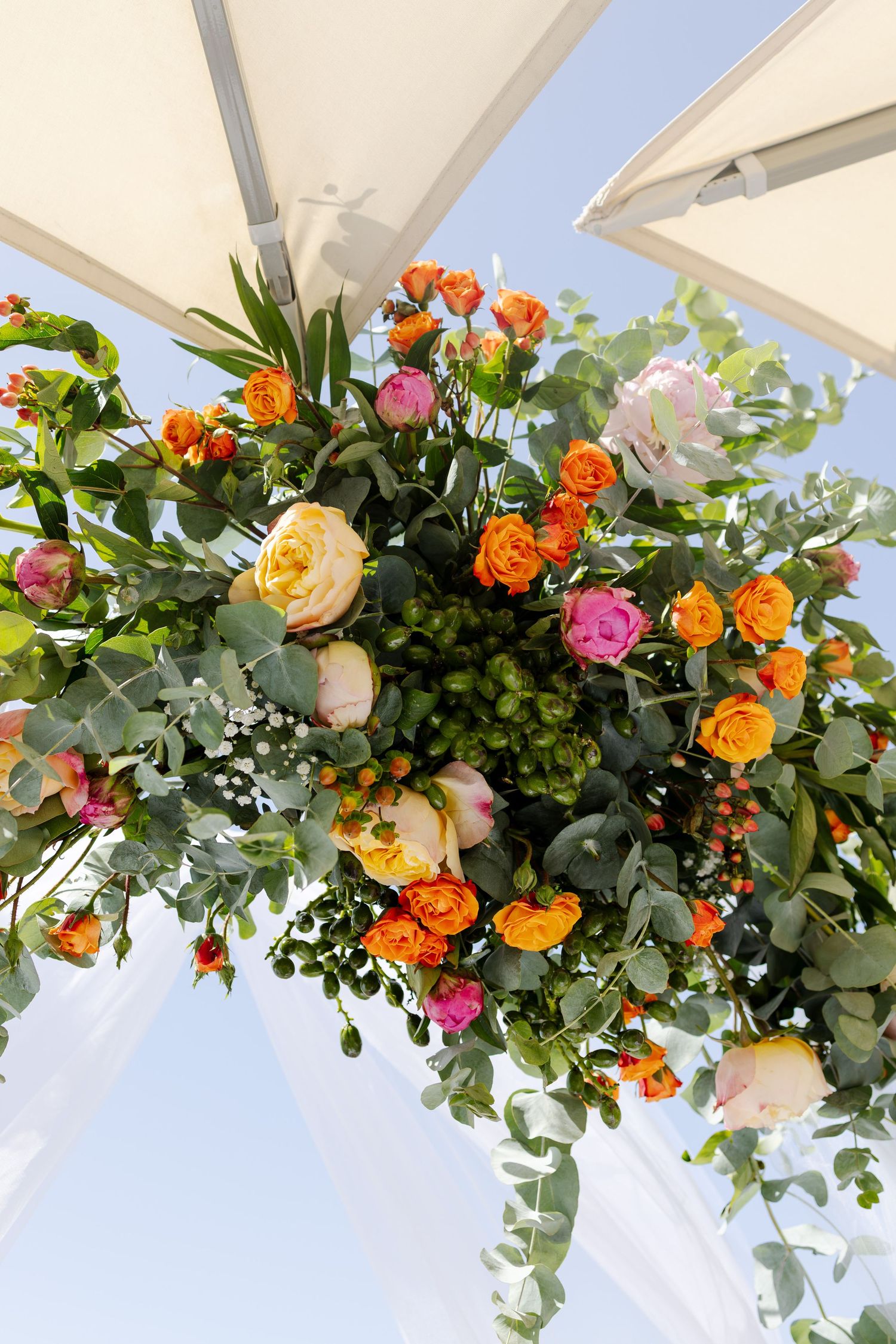 Lush wedding arch decoration featuring vibrant orange, pink, and yellow roses with eucalyptus under a blue sky.