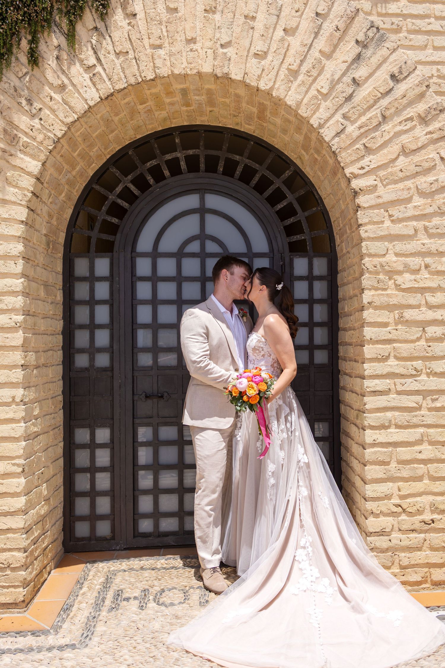 Bride and groom share a romantic kiss under a stone archway holding a colorful bouquet at their wedding.