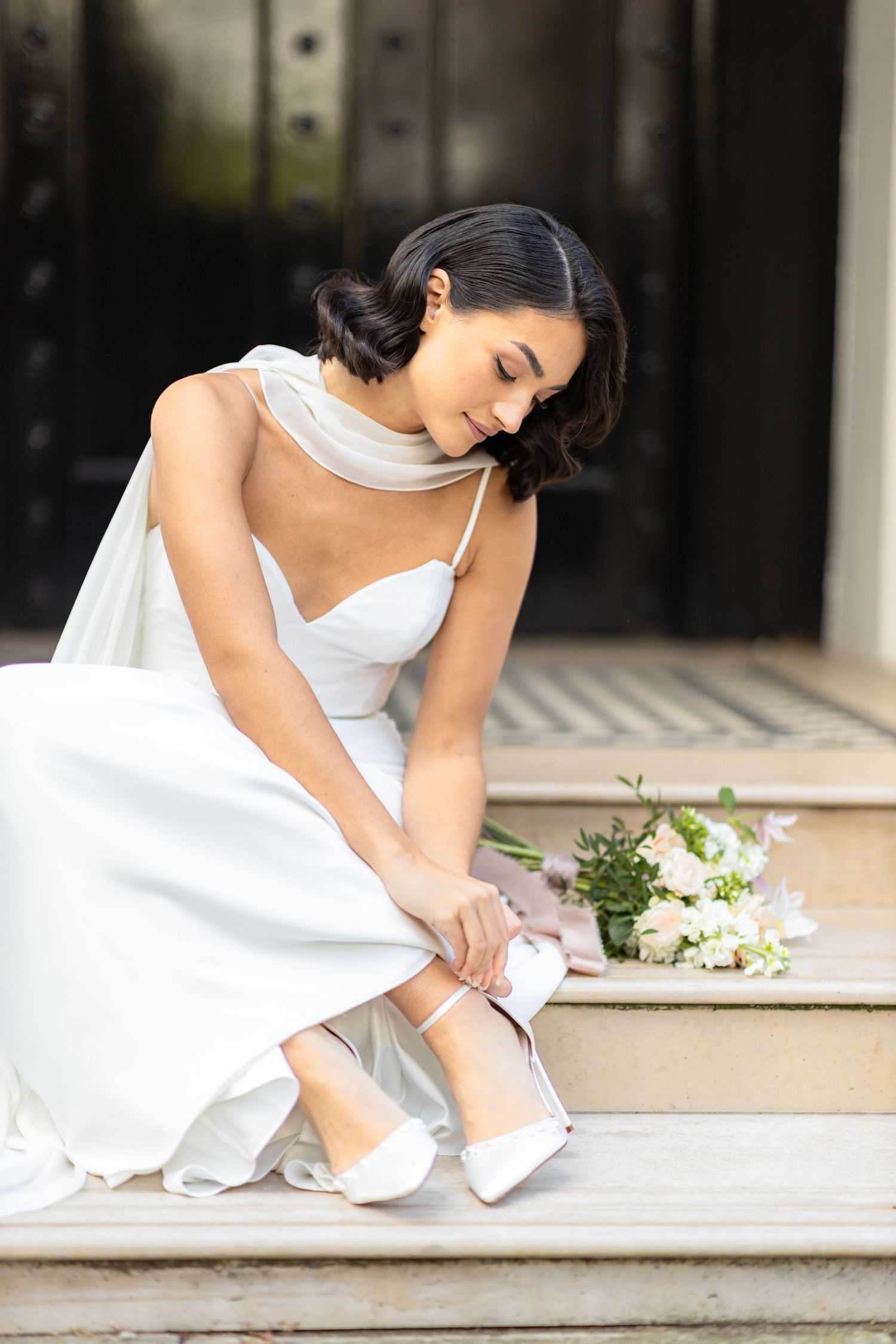 A bride in a white dress adjusts her shoe while sitting on stone steps.