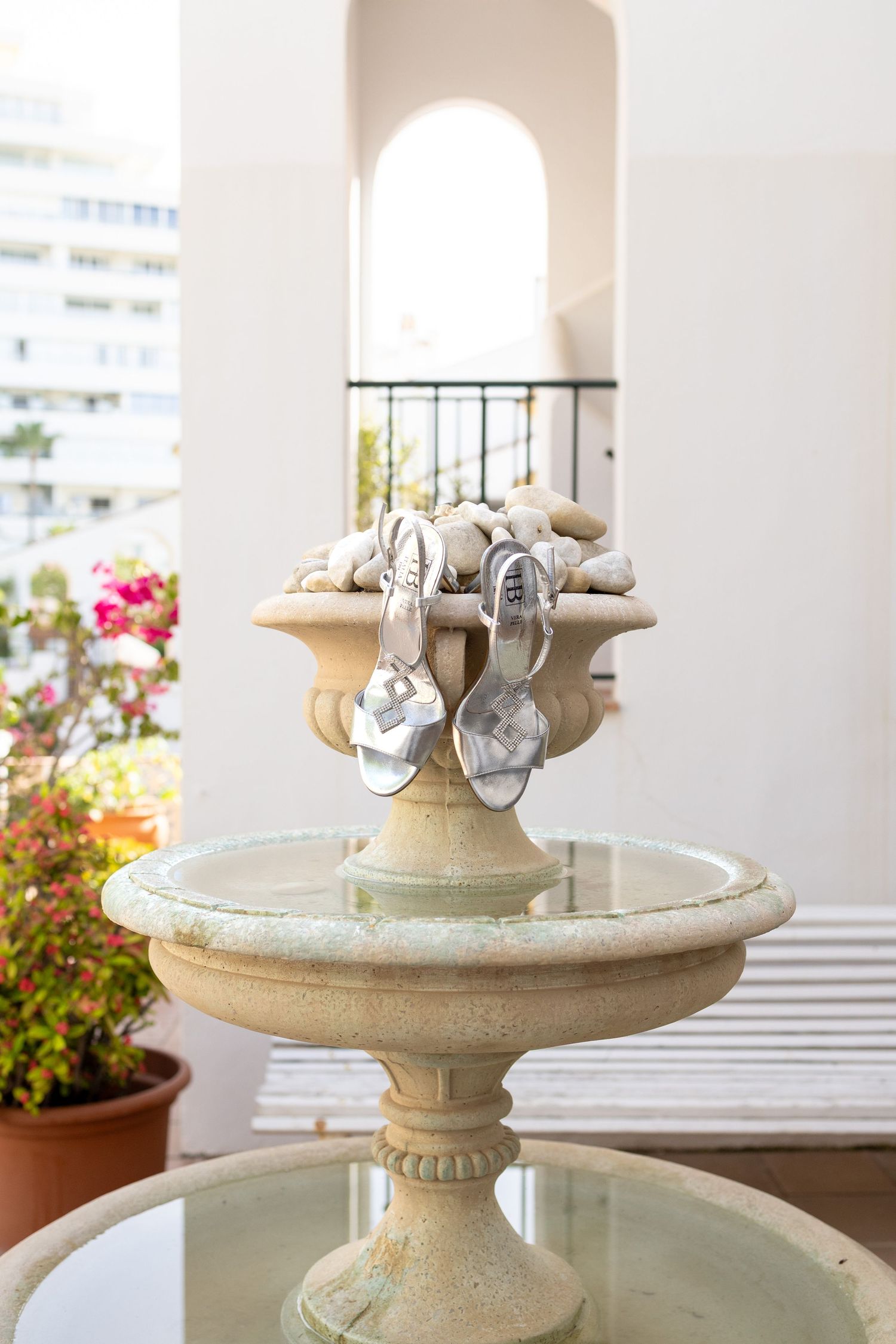 Silver strappy heels displayed on a stone fountain with decorative rocks, set on an elegant outdoor terrace with flowers.