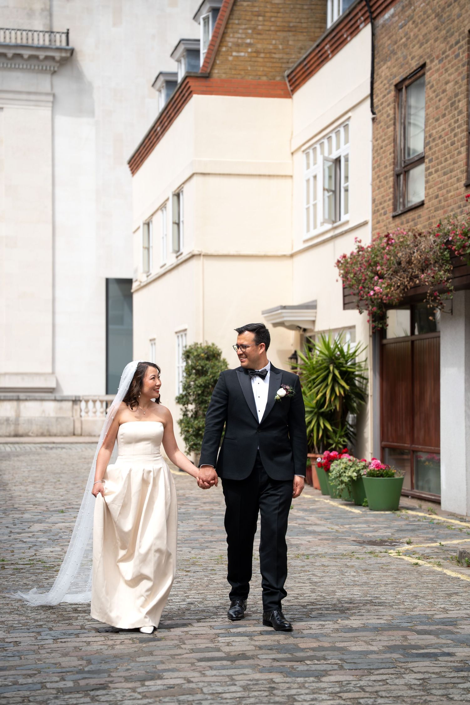 Bride in strapless white gown and groom in black tuxedo walking hand in hand on cobblestone street.