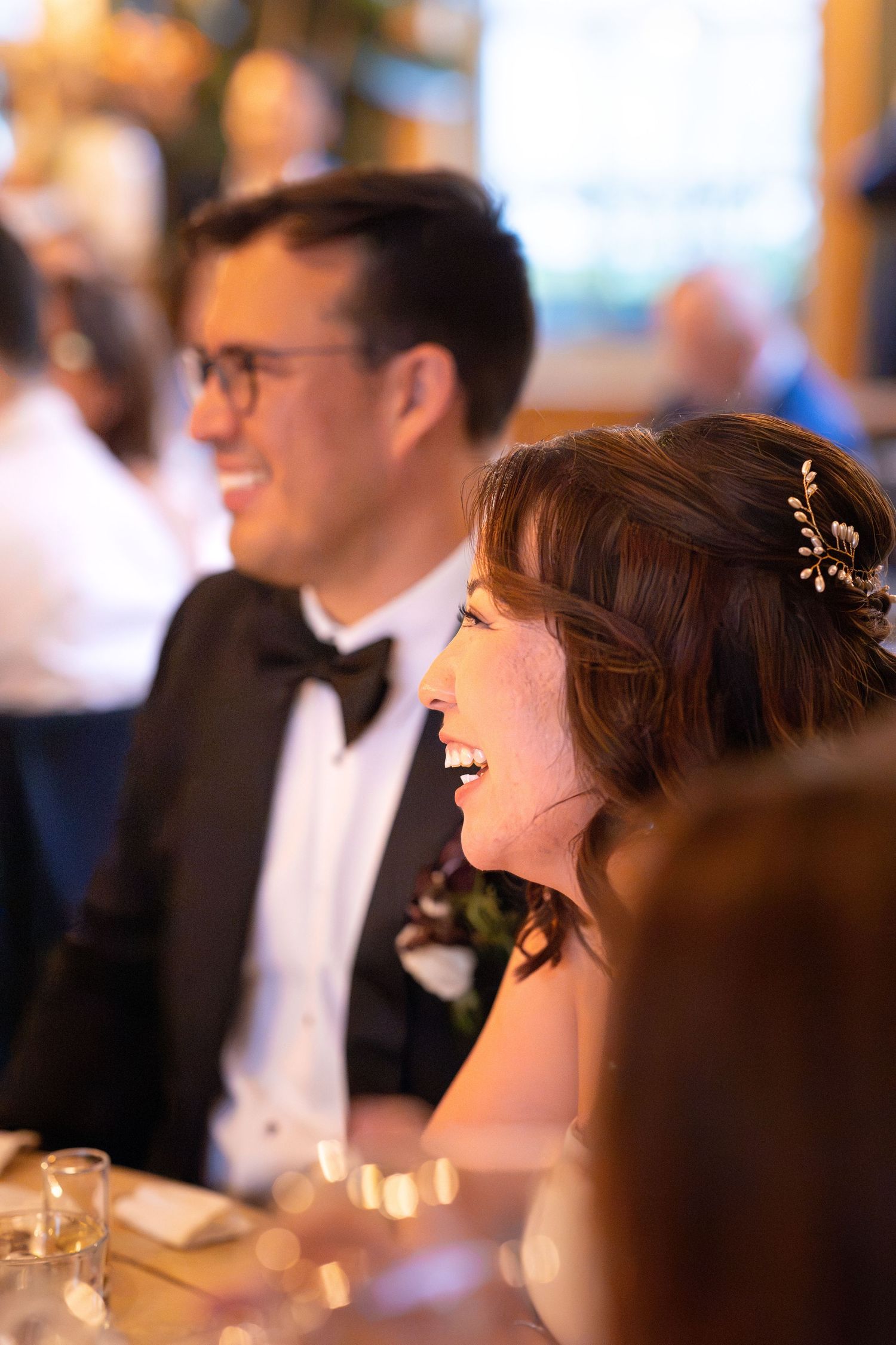 Bride and groom laughing joyfully at wedding reception dinner table, bride wearing floral hair accessory.