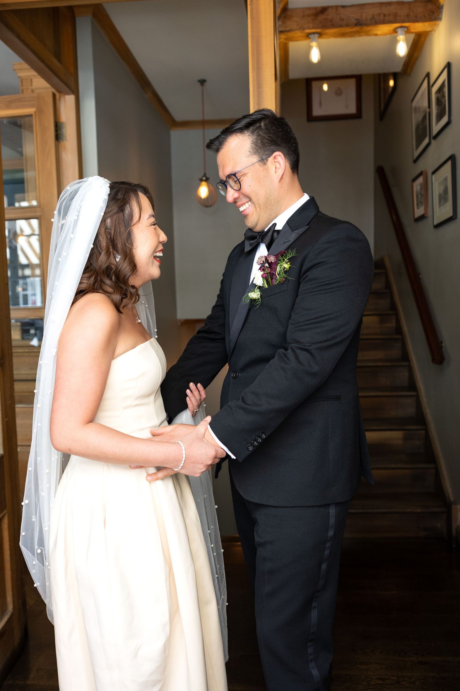 Bride and groom share a joyful first look moment holding hands indoors near a staircase.