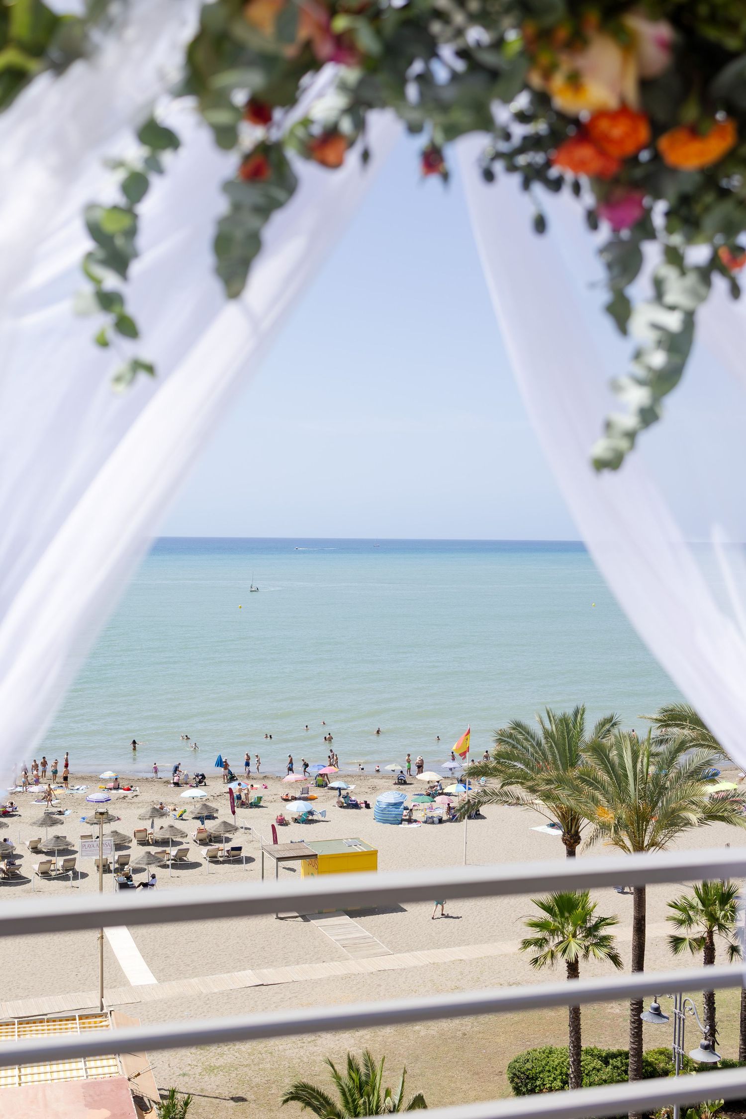 Beach view through white draped wedding arch with tropical flowers, palm trees, and turquoise sea in background.
