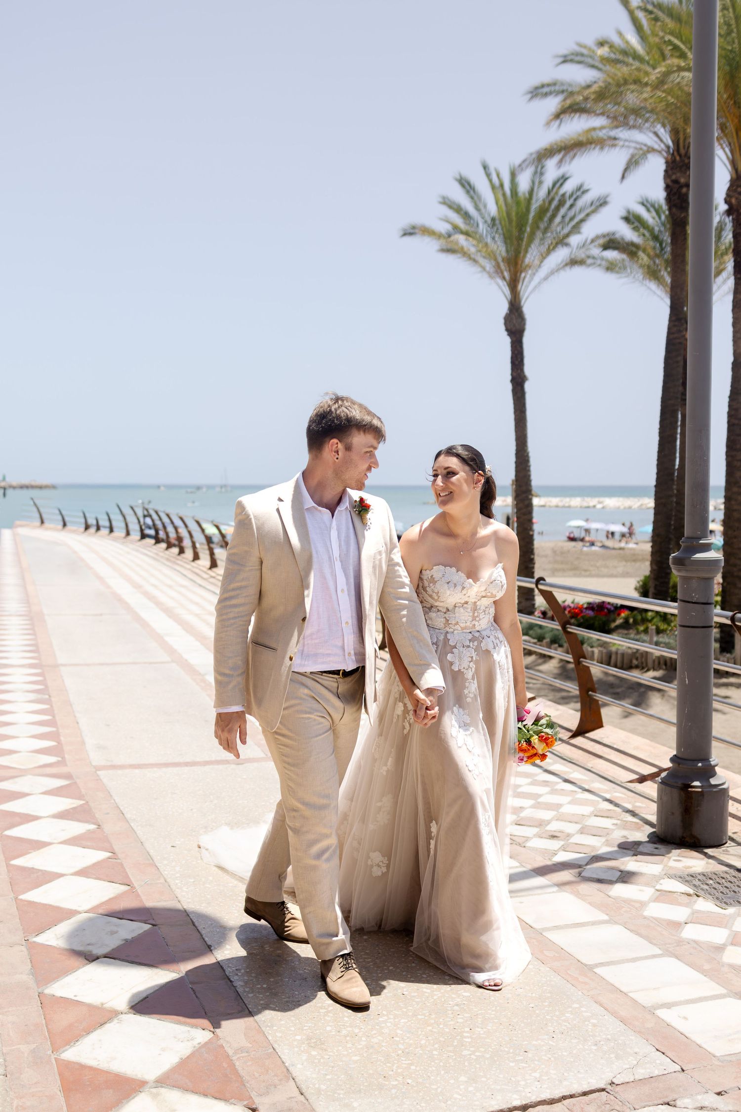 Bride and groom walking hand in hand along a sunny beachside promenade lined with palm trees.