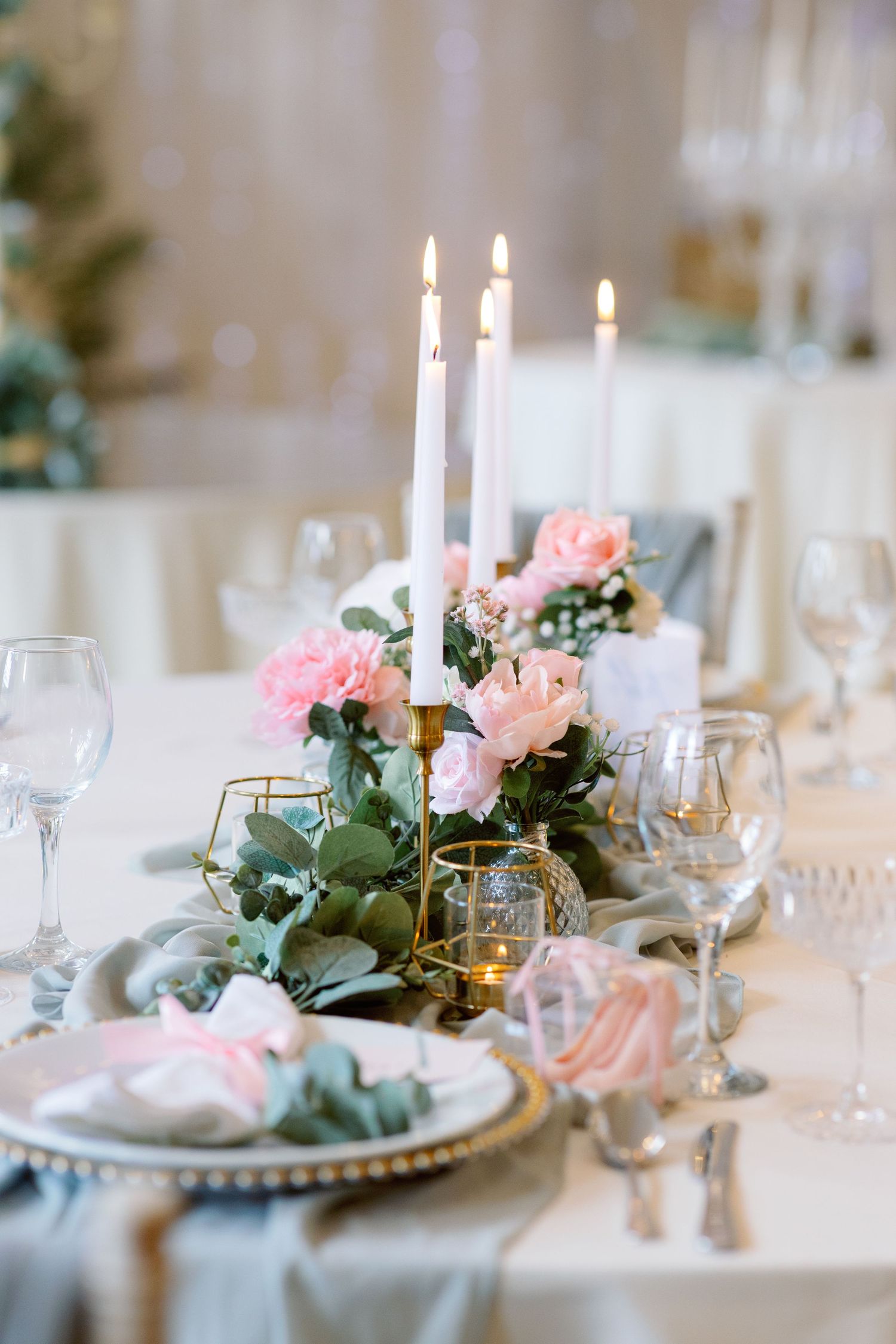 Elegant wedding table setting with white candles, pink roses, and eucalyptus greenery on a white tablecloth.