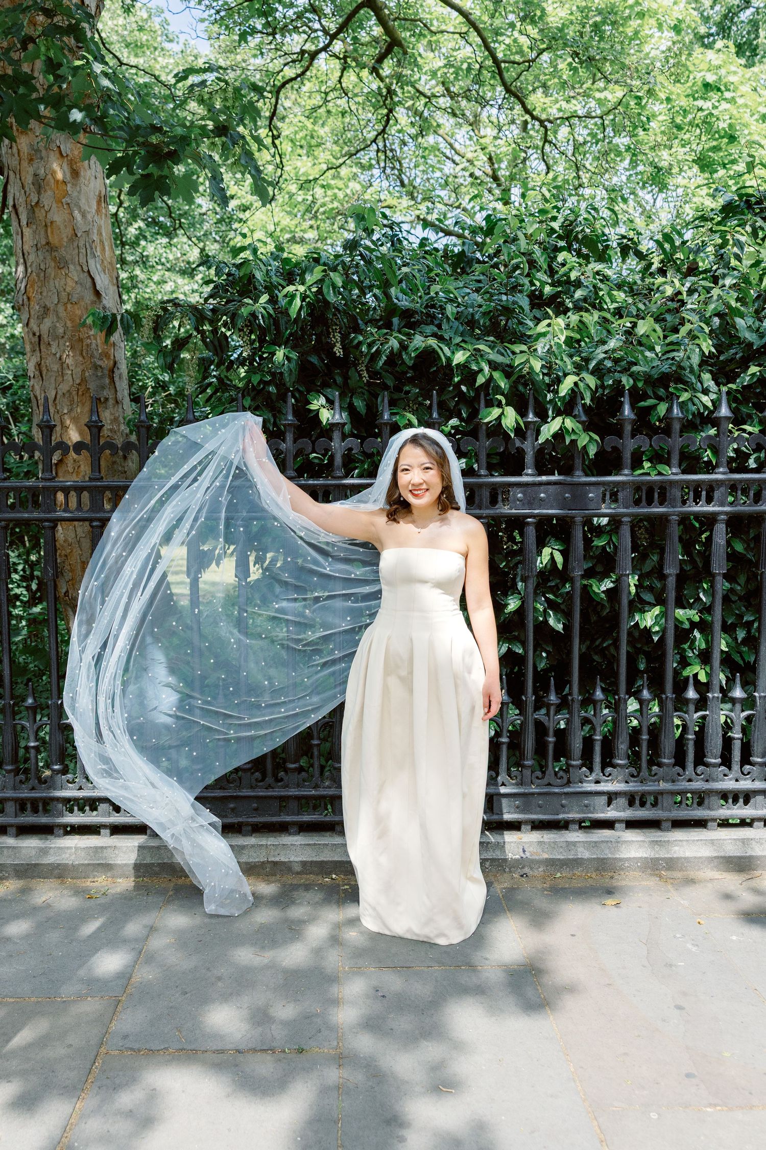 Bride in strapless white gown with flowing veil caught in breeze beside iron fence.