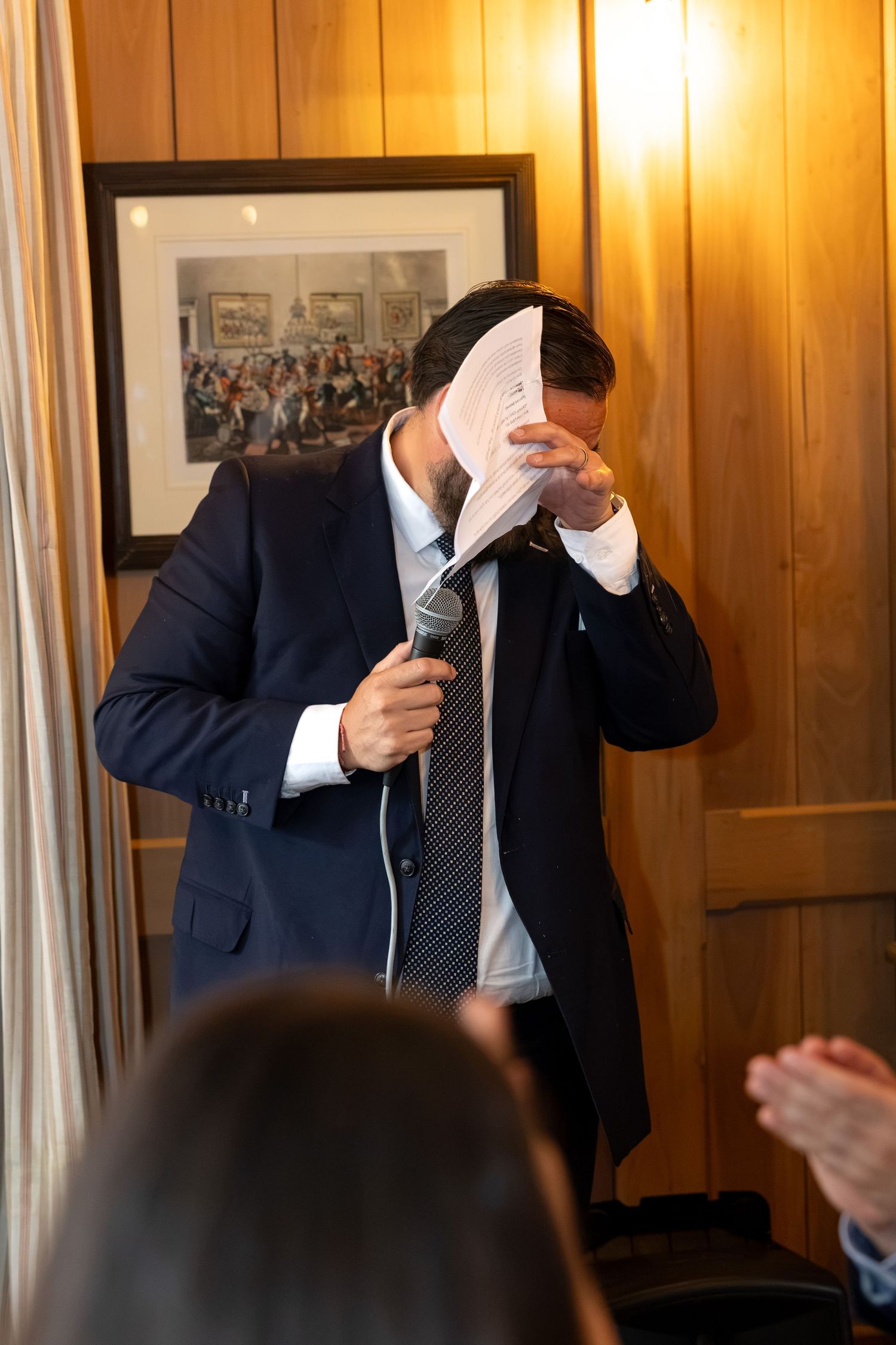 Emotional man in suit covering face while giving wedding speech with microphone.