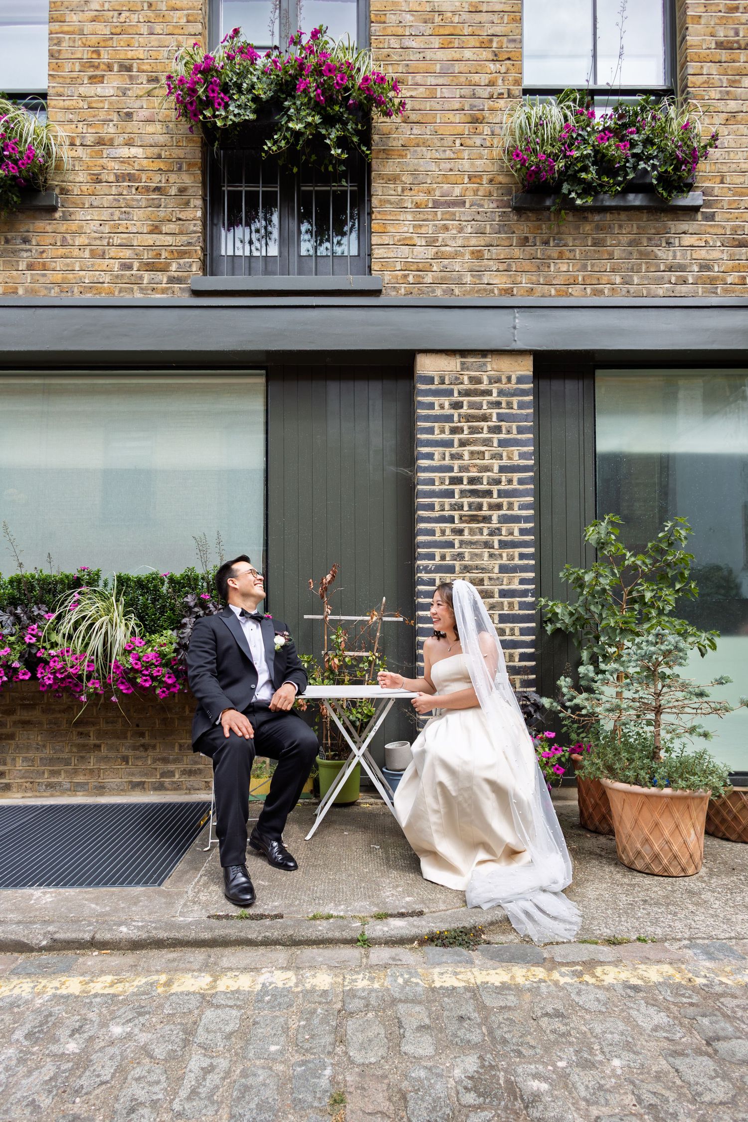 Bride and groom sit at outdoor cafe table on cobblestone street beside brick building adorned with colorful flower boxes.