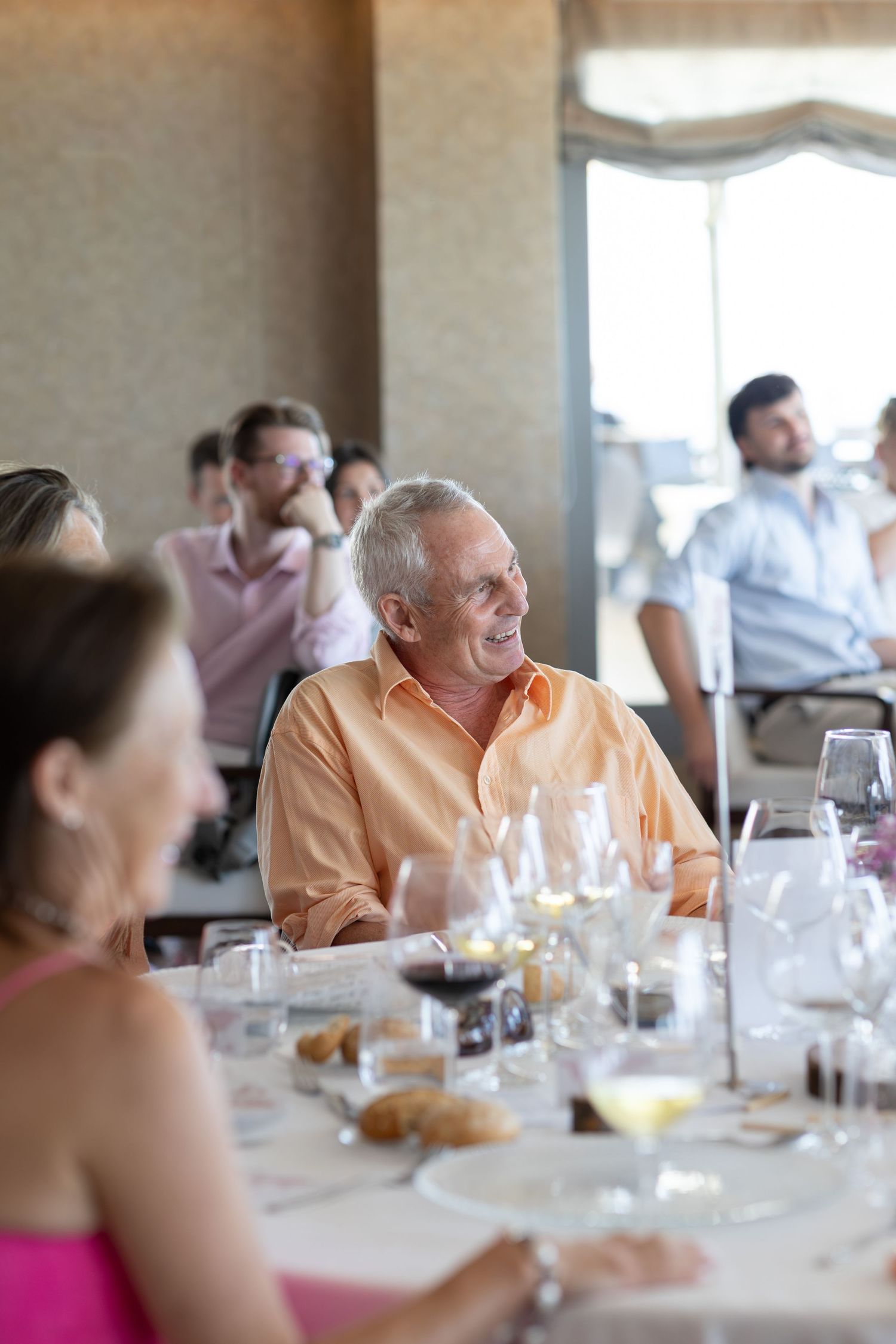 Smiling senior man in orange shirt enjoying wedding reception dinner with guests at elegantly set table.