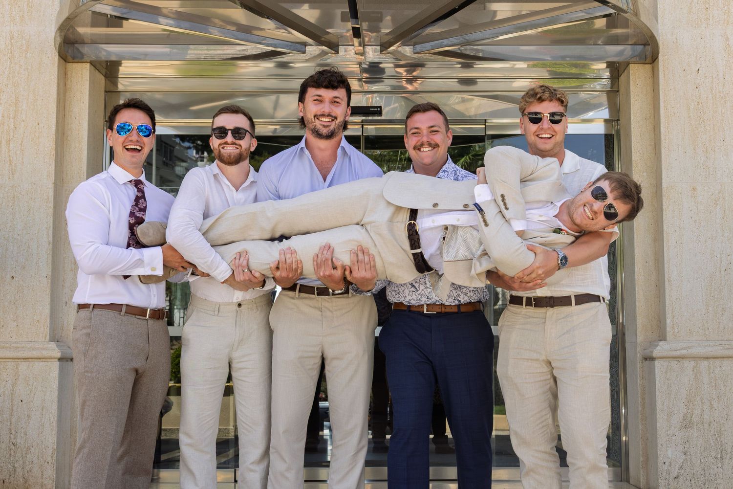 A group of five groomsmen in tan suits playfully lift one of their friends outside a venue.