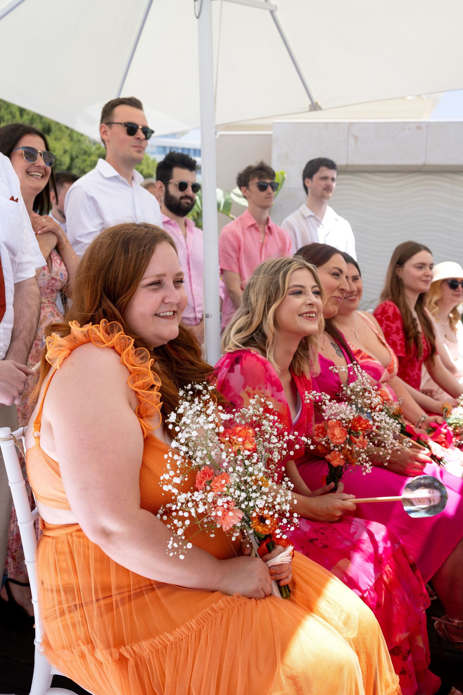 Wedding guests in vibrant orange and pink dresses hold floral bouquets while seated at an outdoor ceremony.