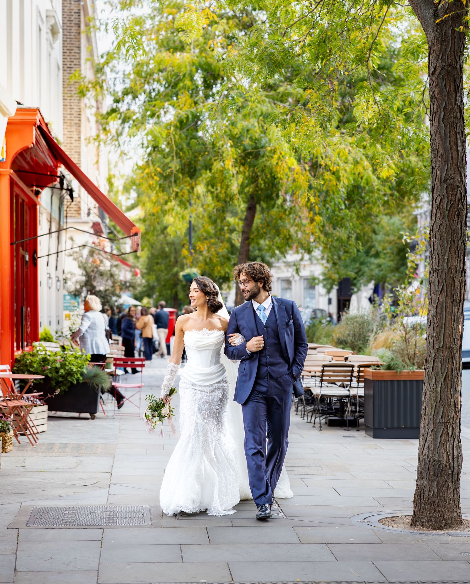 A couple in wedding attire walks together along a tree-lined pathway.