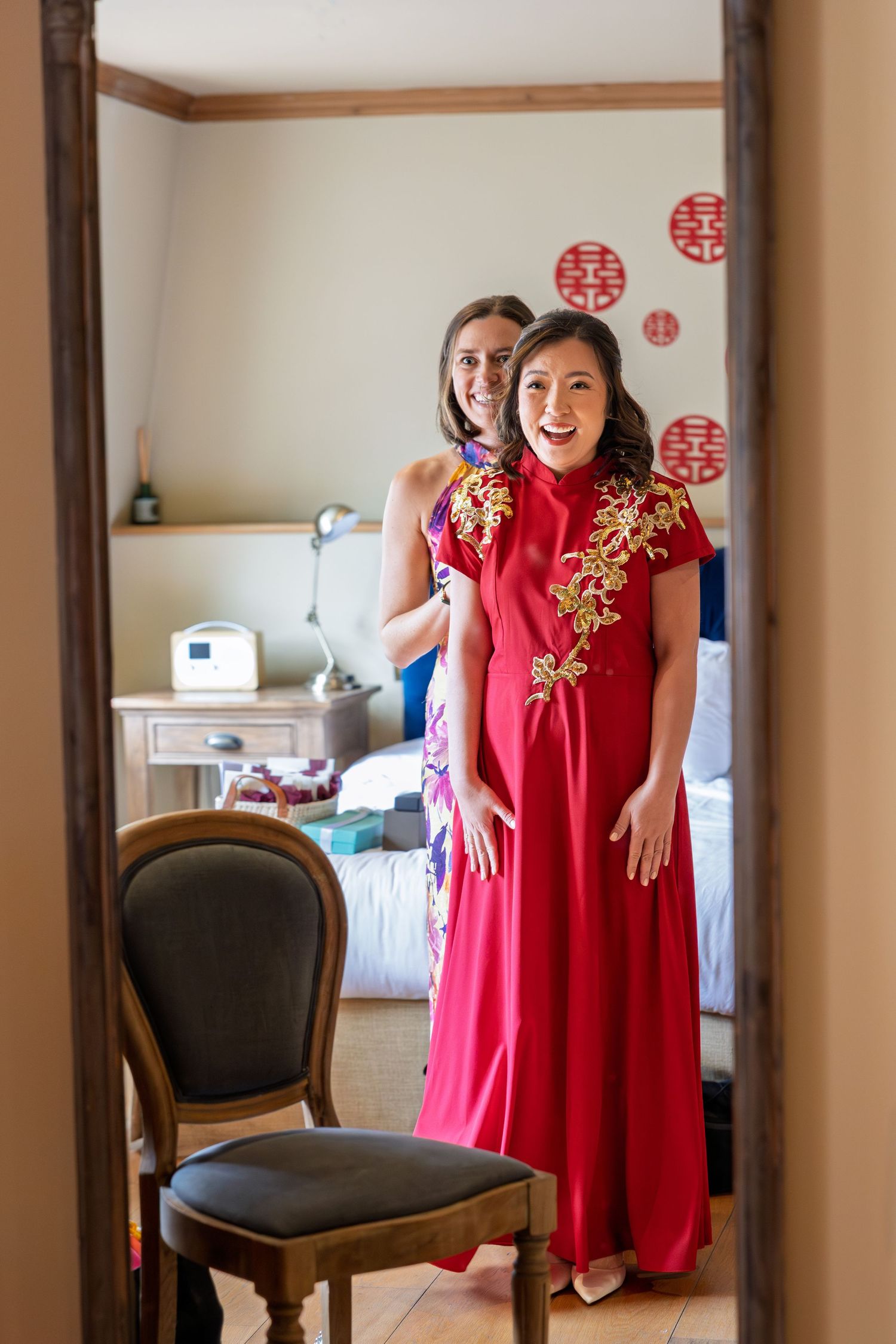 Bride in red Chinese qipao with gold floral embroidery smiling in mirror reflection during wedding preparations.