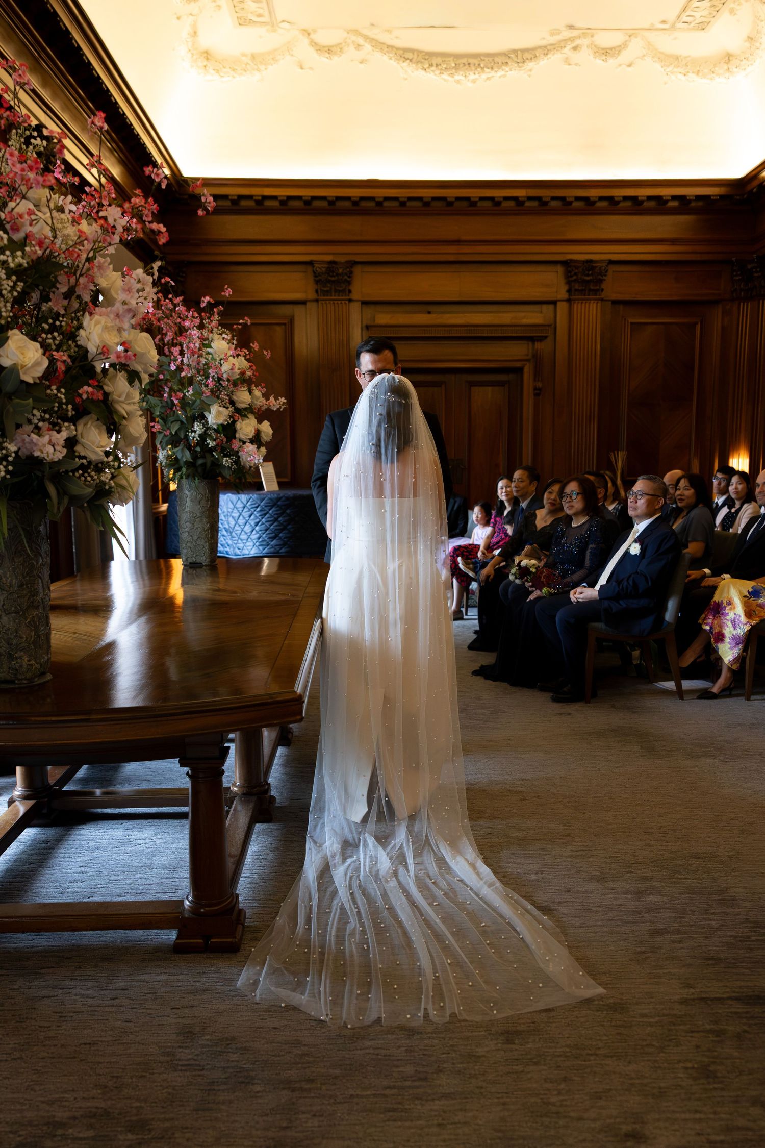 Bride in long veil stands at altar with groom before seated wedding guests in elegant wood-paneled room.