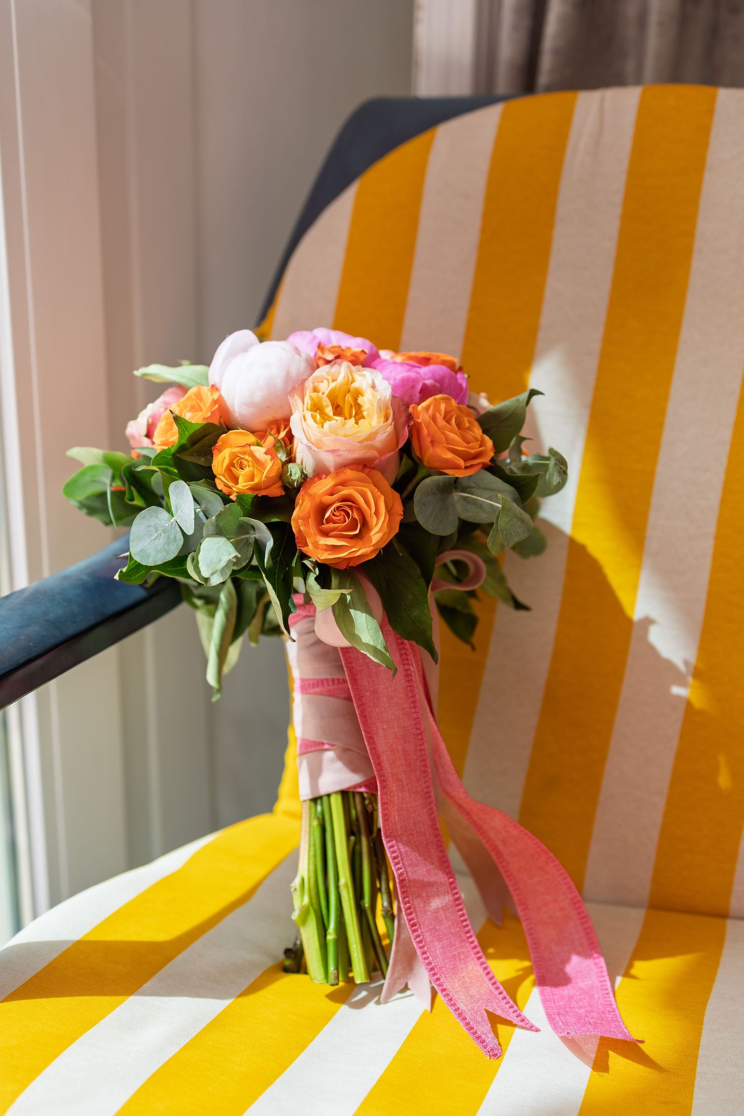 A vibrant bridal bouquet of orange and pink roses with ribbons rests on a yellow striped chair.