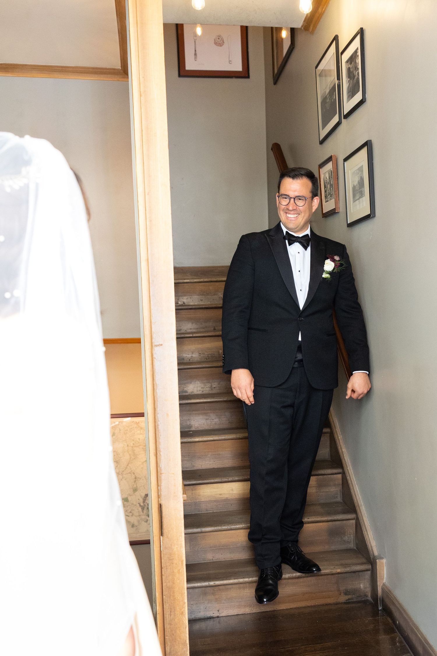 Smiling groom in black tuxedo stands at bottom of staircase as bride in veil approaches.