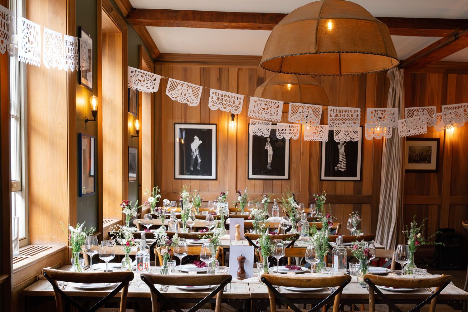 Elegant wedding reception room with long tables, floral centerpieces, papel picado banners, and warm lighting.