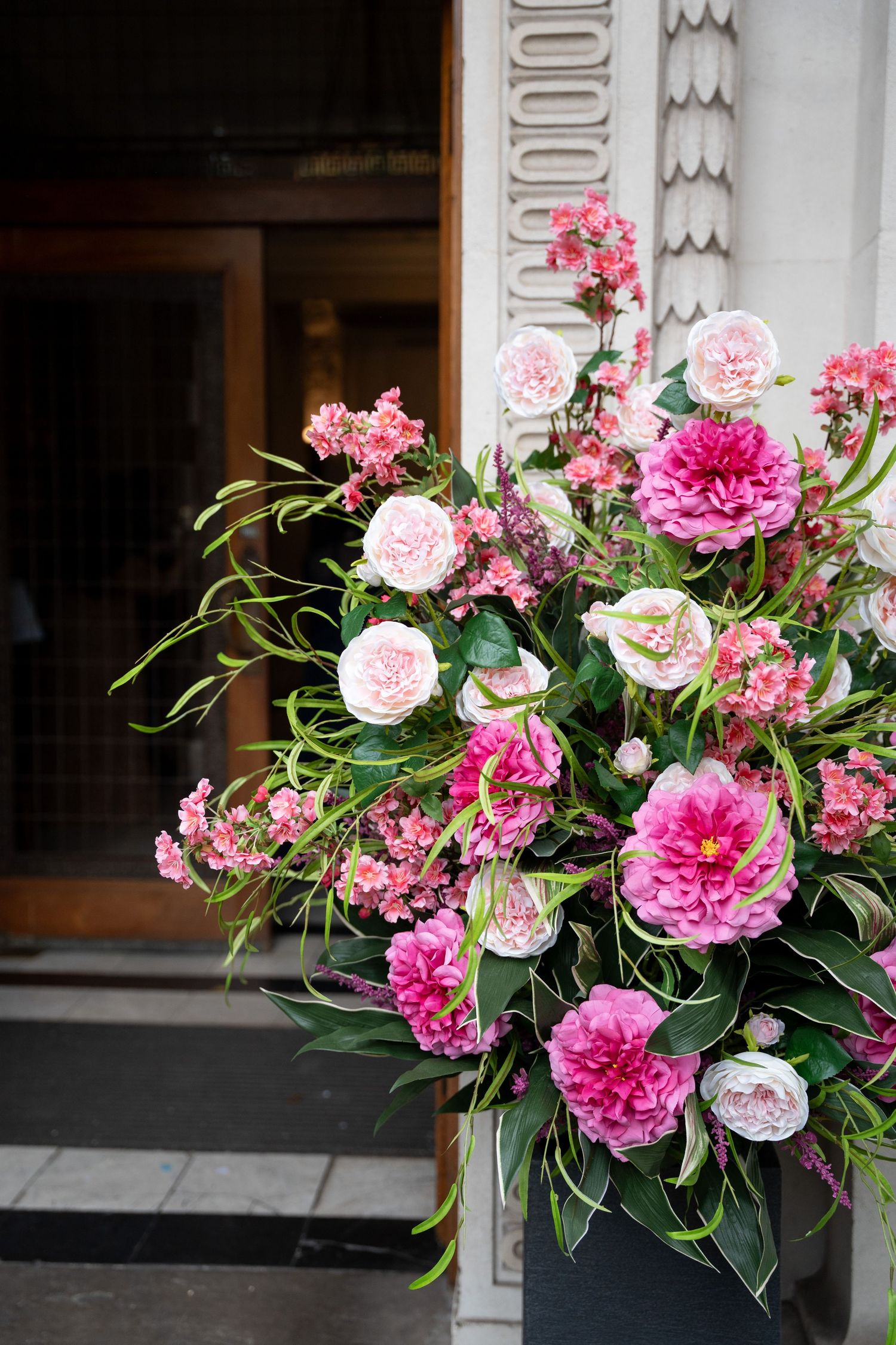 Lush pink peony and rose floral arrangement outside an elegant building entrance.