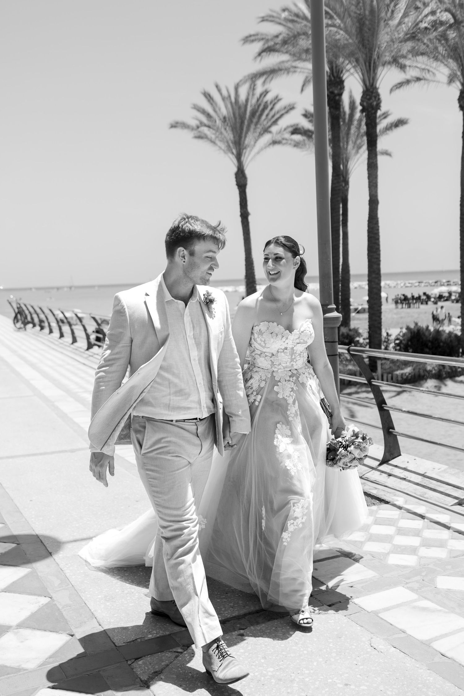 Black and white photo of a bride and groom walking along a sunny palm-lined promenade near the beach.