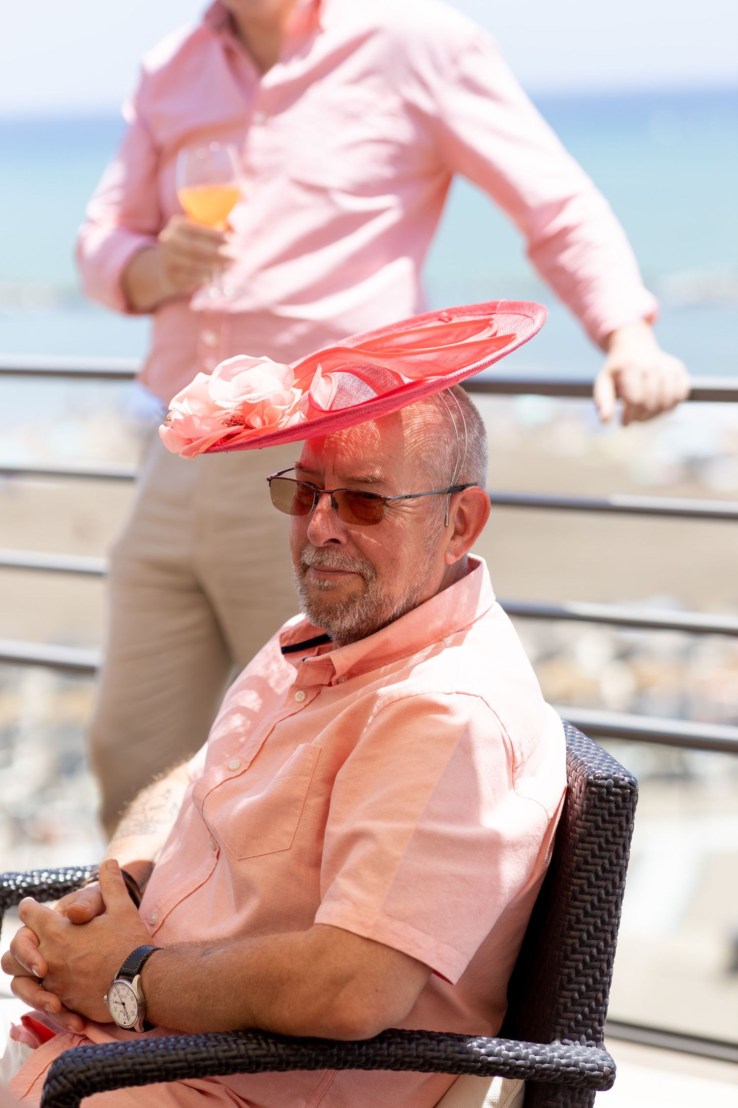 Elderly man wearing pink shirt and large red fascinator hat seated at a sunny beachside venue.
