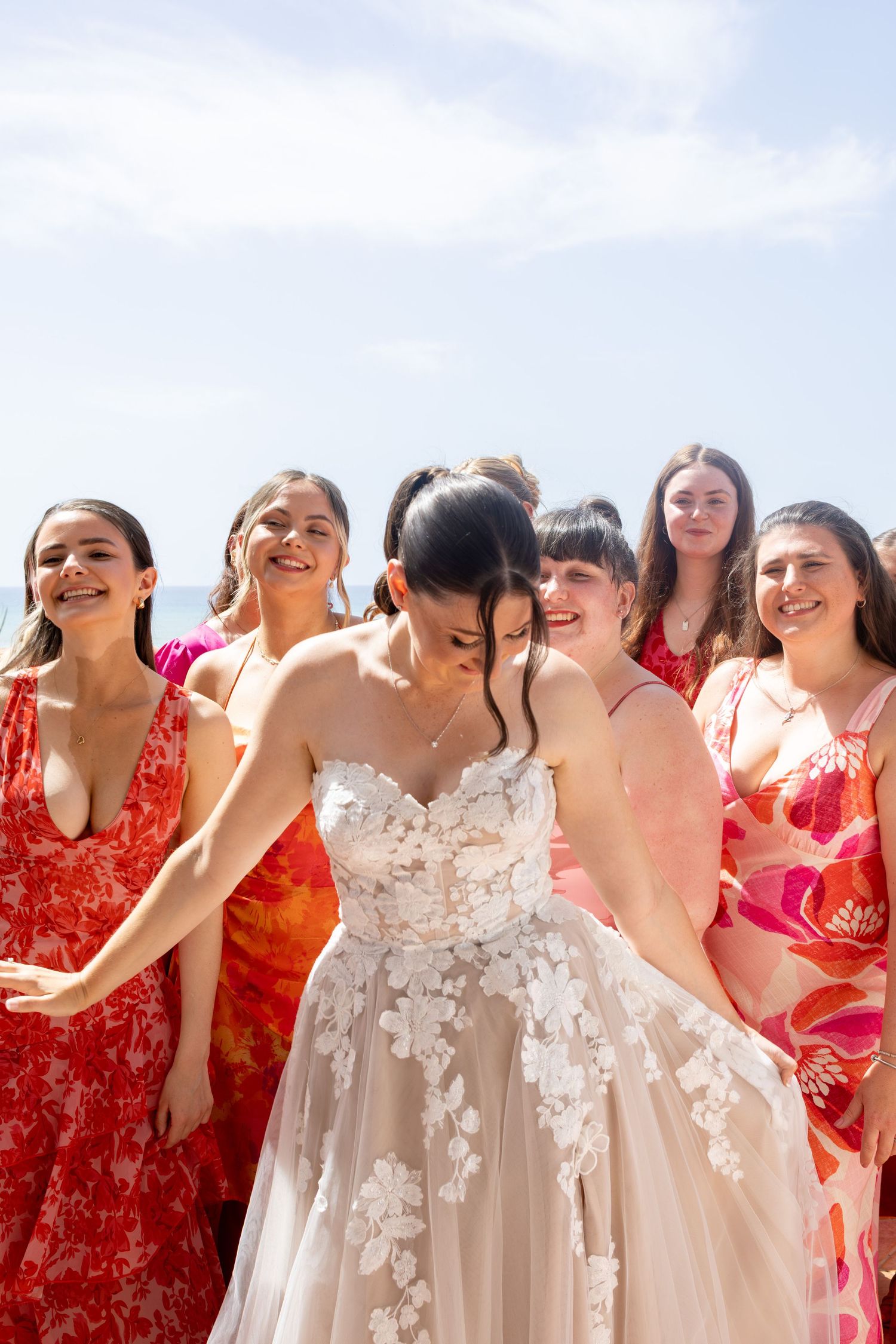 Bride in floral wedding gown laughing with bridesmaids in red and pink dresses at a sunny beach wedding.