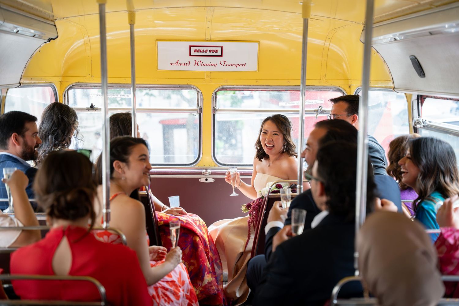 A bride laughs and chats with guests while riding inside a vintage yellow double-decker wedding party bus.