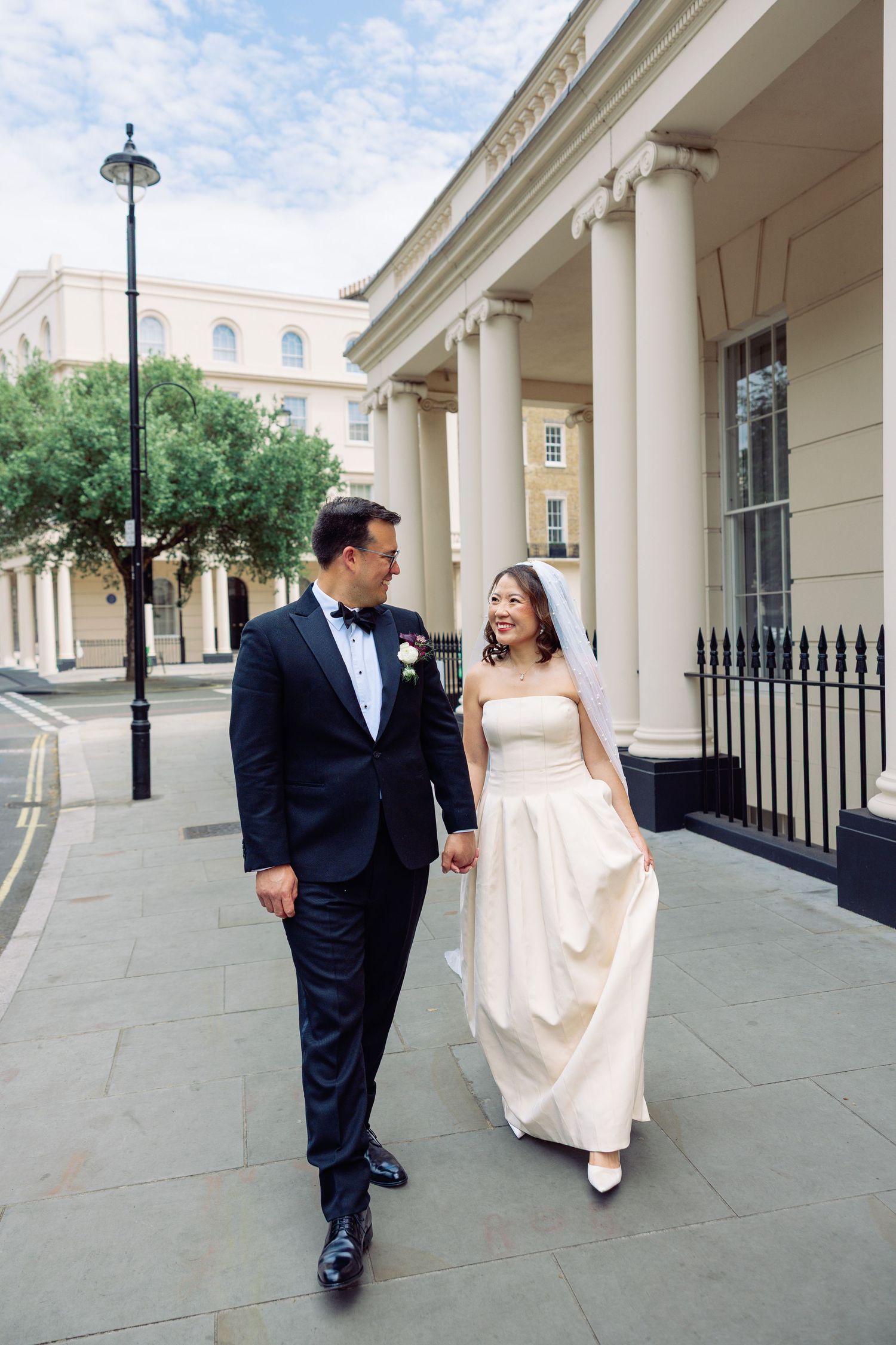 Newlyweds walking hand in hand along a sunny London street in wedding attire.