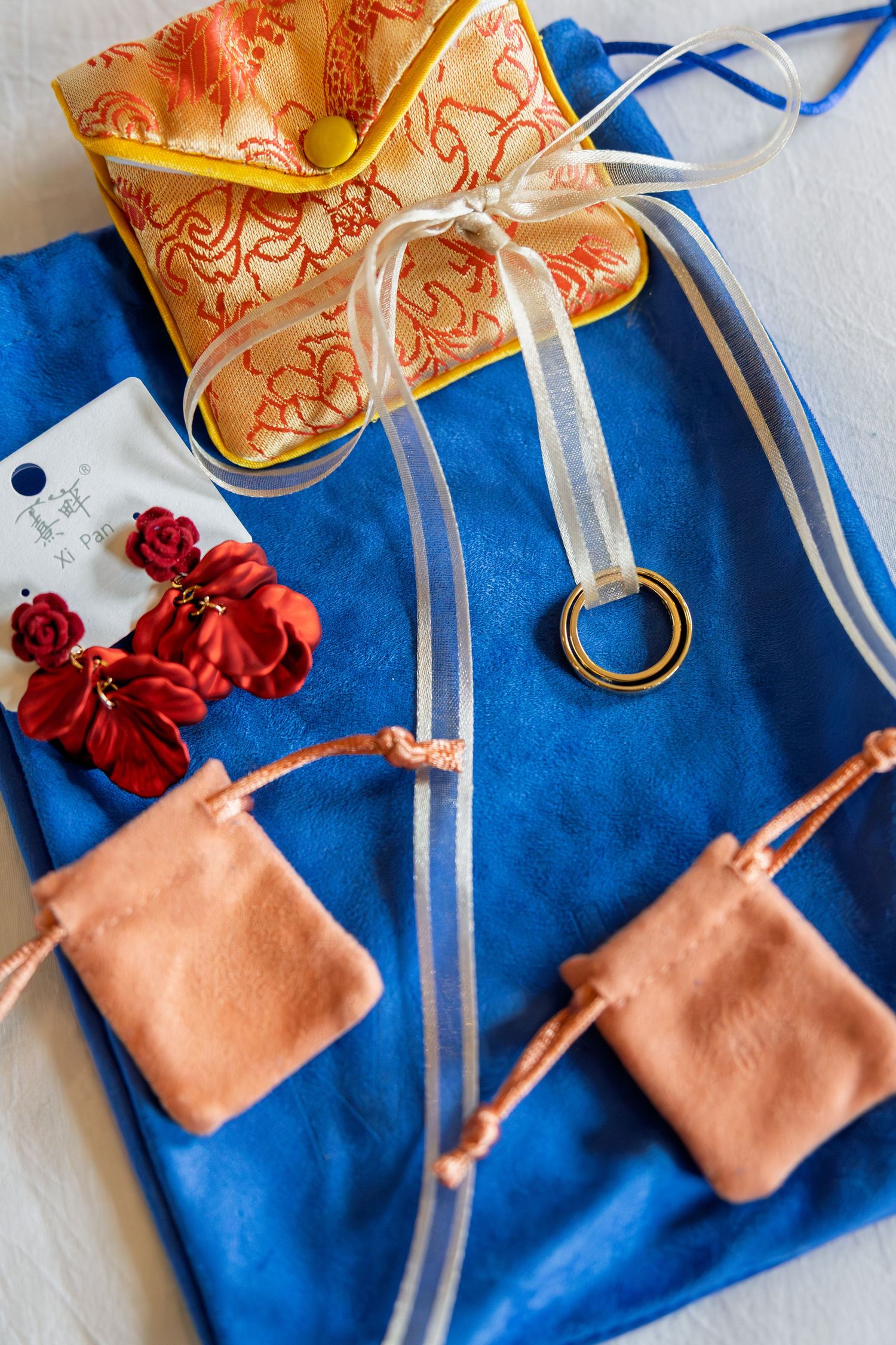 Blue suede jewelry roll with red floral earrings, gold ring, peach pouches, and ornate Chinese brocade pouch with ribbon.