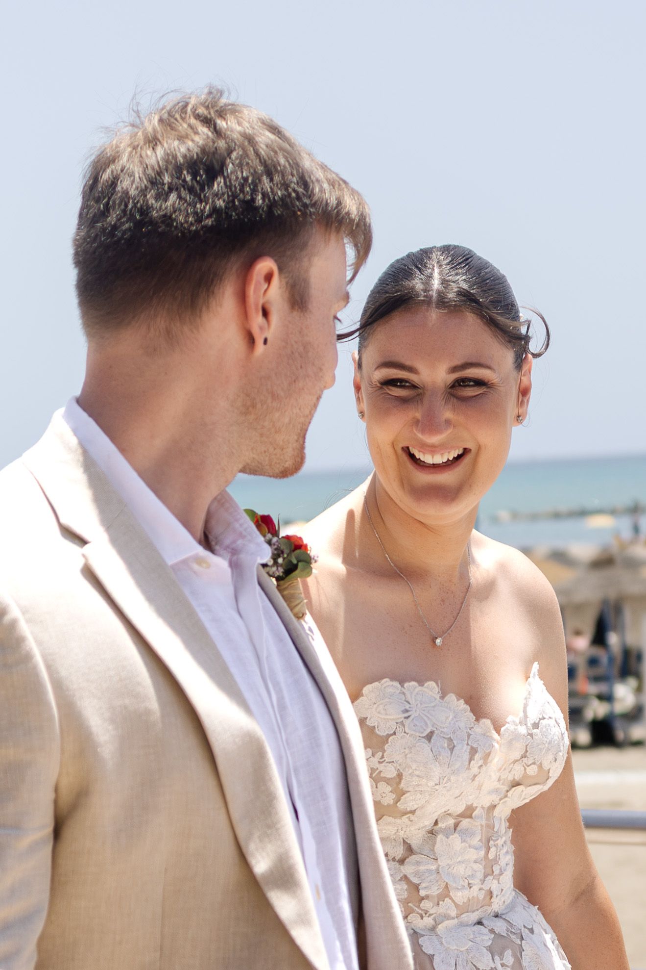 Happy bride in lace gown laughing with groom in beige suit at sunny beach wedding ceremony.