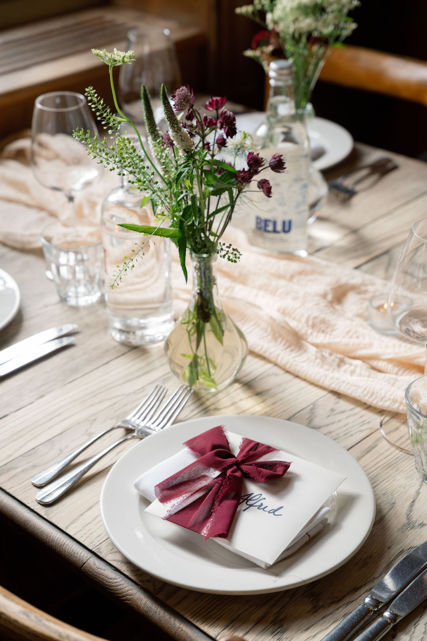 Elegant wedding table setting with burgundy bow napkin, wildflower centerpiece, and Belvu water bottle.