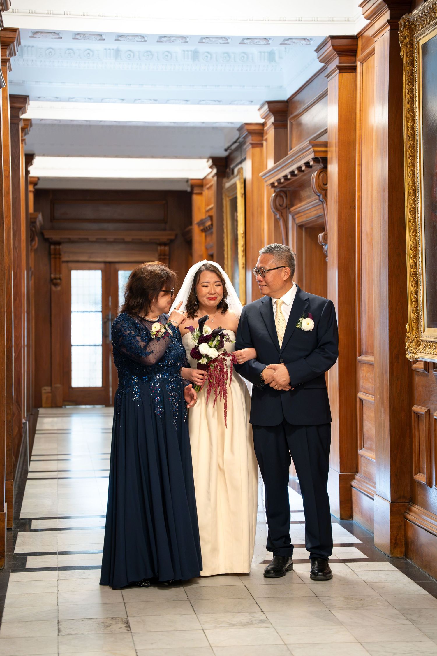 Bride in white gown walks with parents through elegant wood-paneled wedding venue hallway.