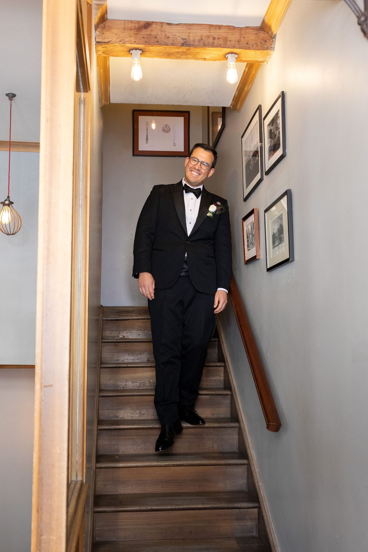 Groom in black tuxedo with bow tie and boutonniere descending wooden staircase at wedding venue.