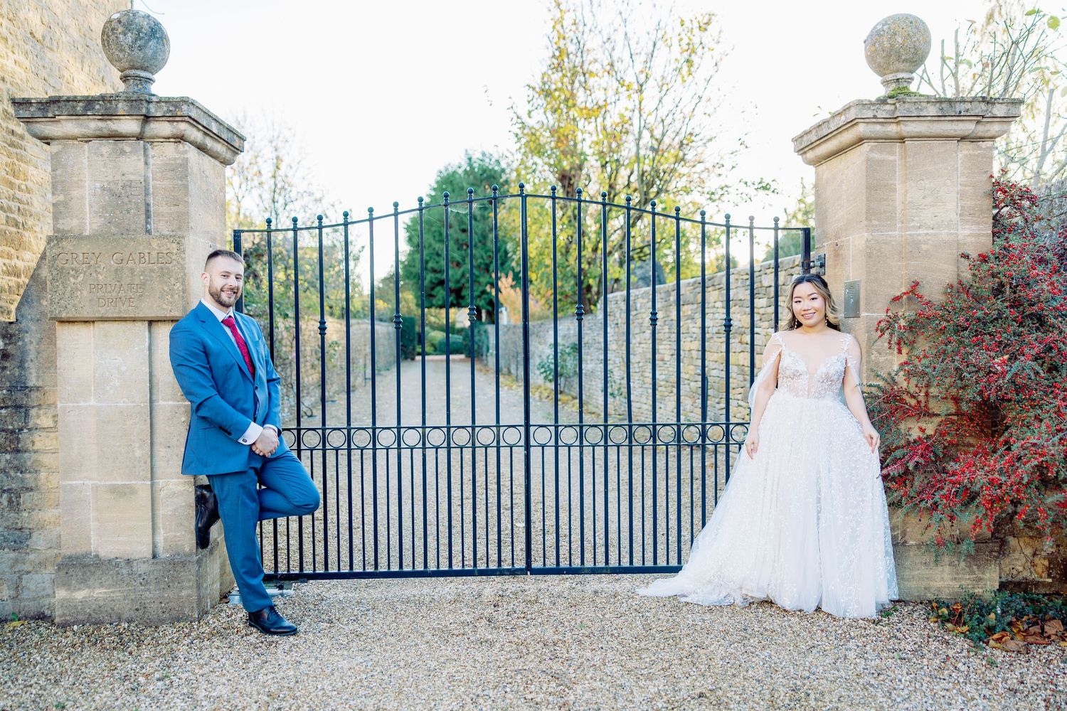 Wedding couple poses by ornate stone gates and autumn foliage.