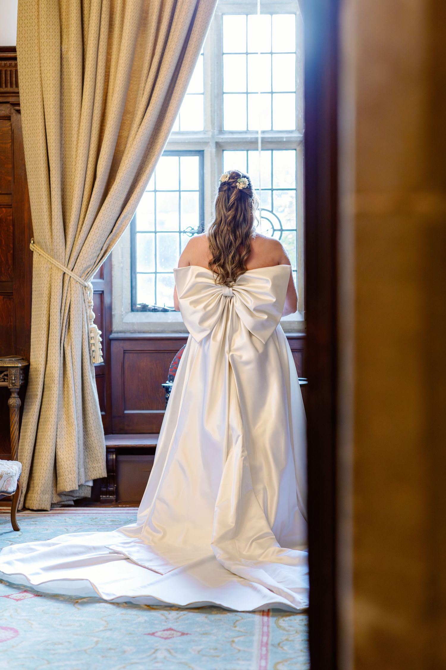 A white wedding dress with a dramatic bow detail photographed in an elegant room with cream curtains and natural light.