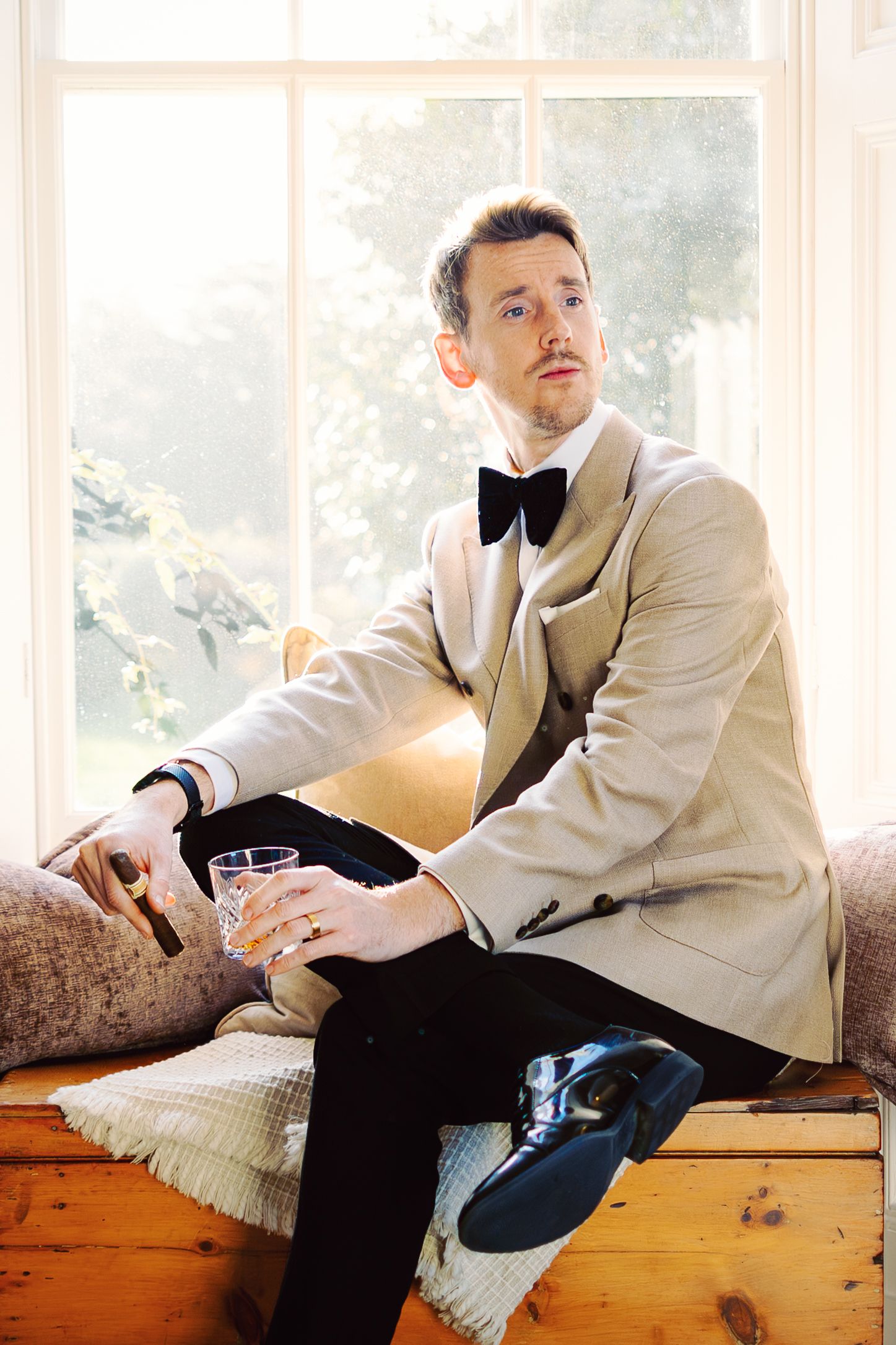Elegant groom in cream tuxedo jacket and bow tie poses by window with cocktail glass.