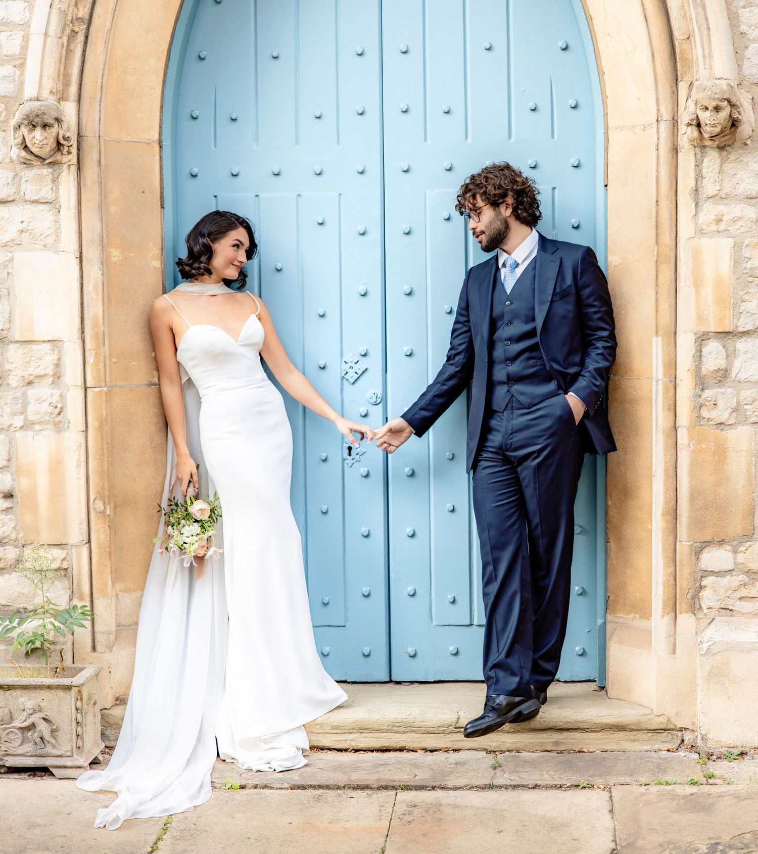 Elegant wedding couple poses against a light blue vintage door and stone wall backdrop.