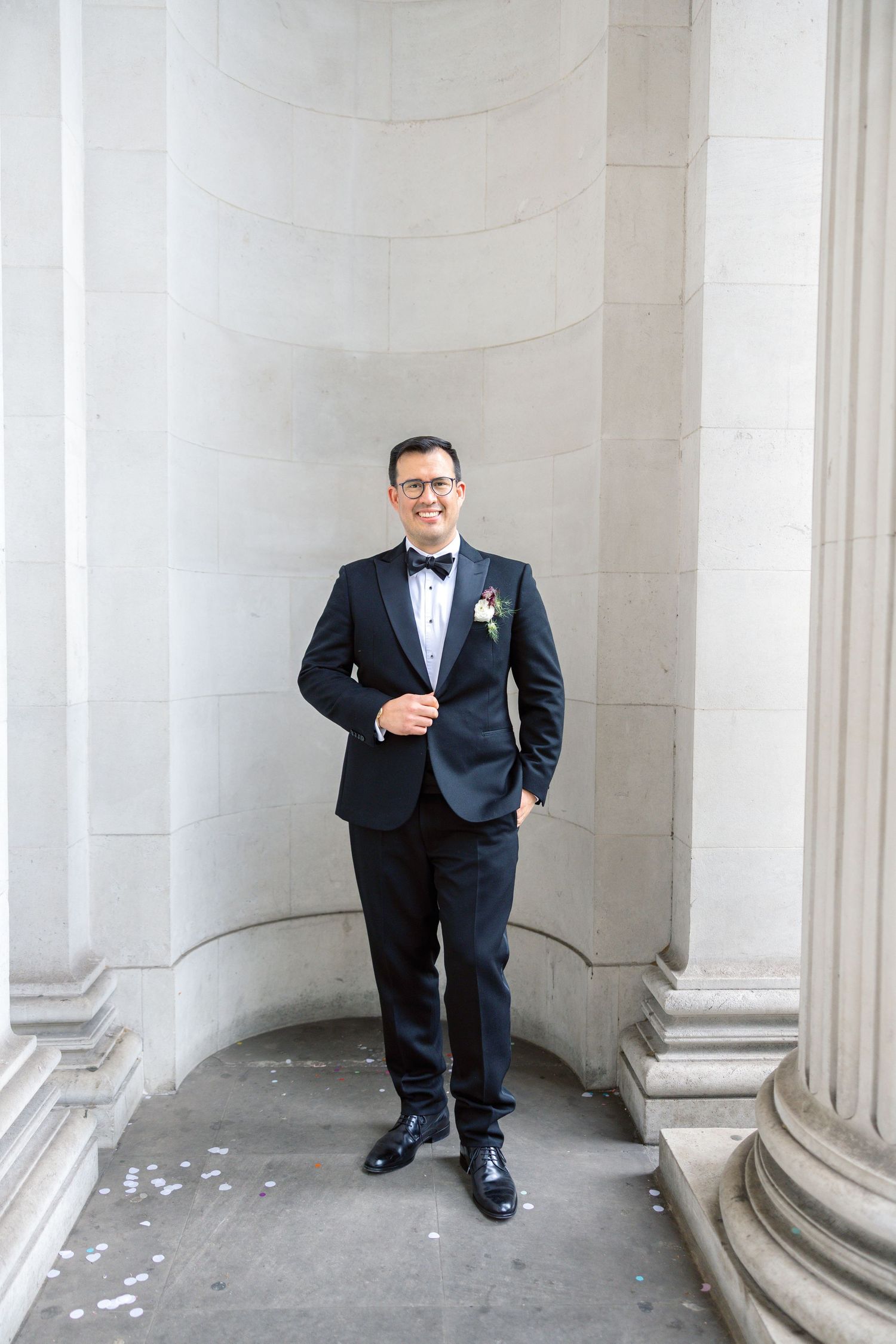 A groom in a black tuxedo with bow tie and boutonniere stands confidently between stone columns.