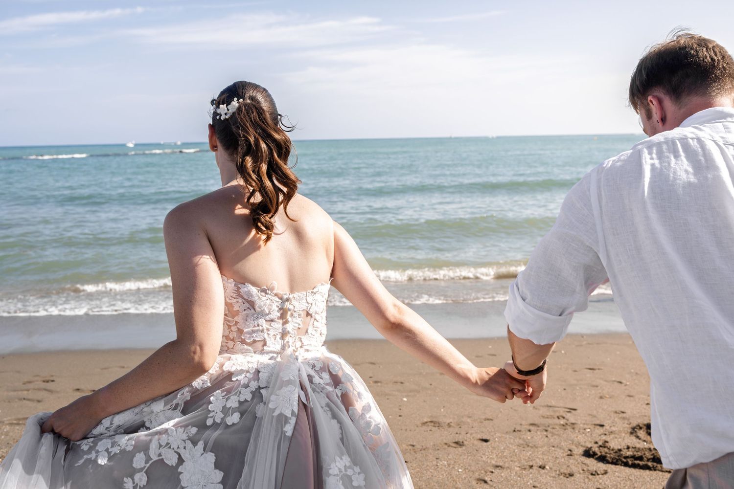 Bride in floral wedding gown holding hands with groom while facing the ocean on sandy beach.