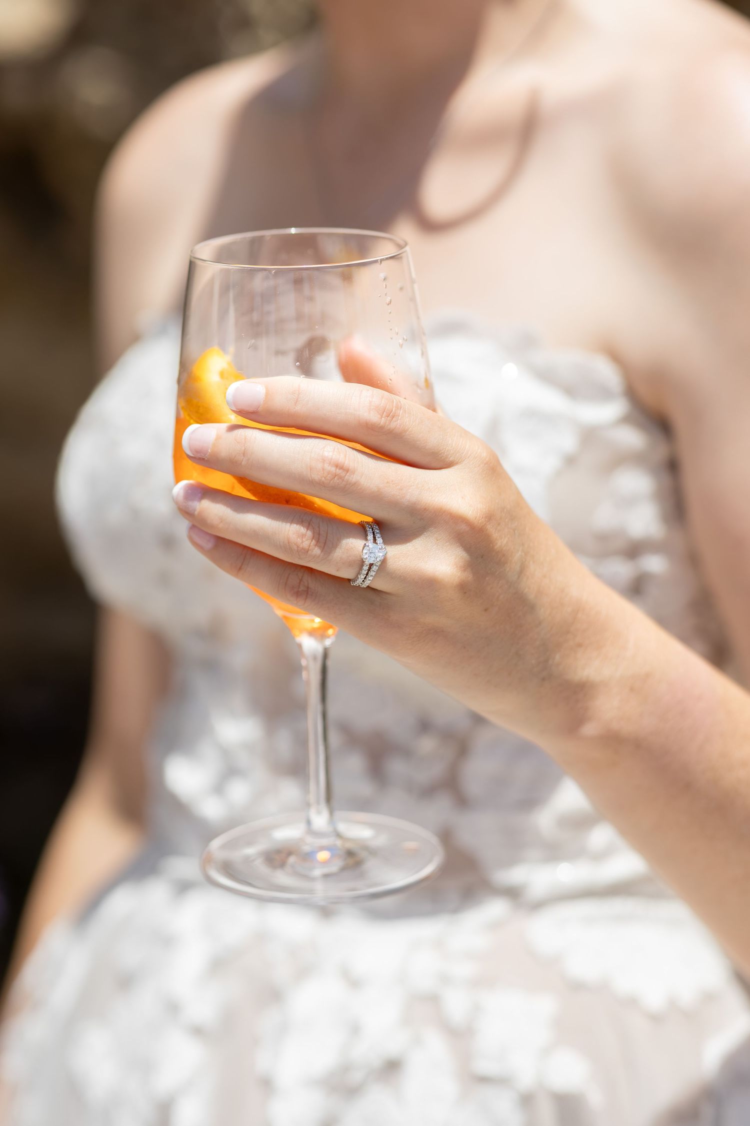 Close-up of a bride holding a wine glass with an orange garnish, wearing a diamond engagement ring.