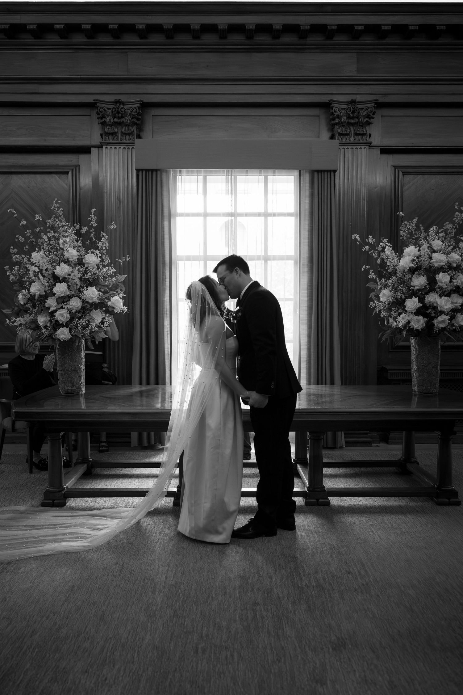 Black and white photo of bride and groom sharing first kiss before tall window flanked by floral arrangements.
