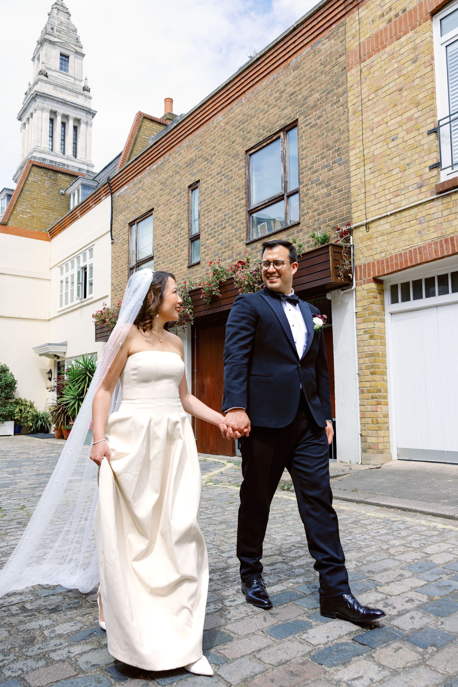 Bride and groom walk hand in hand on cobblestone street with historic London architecture.