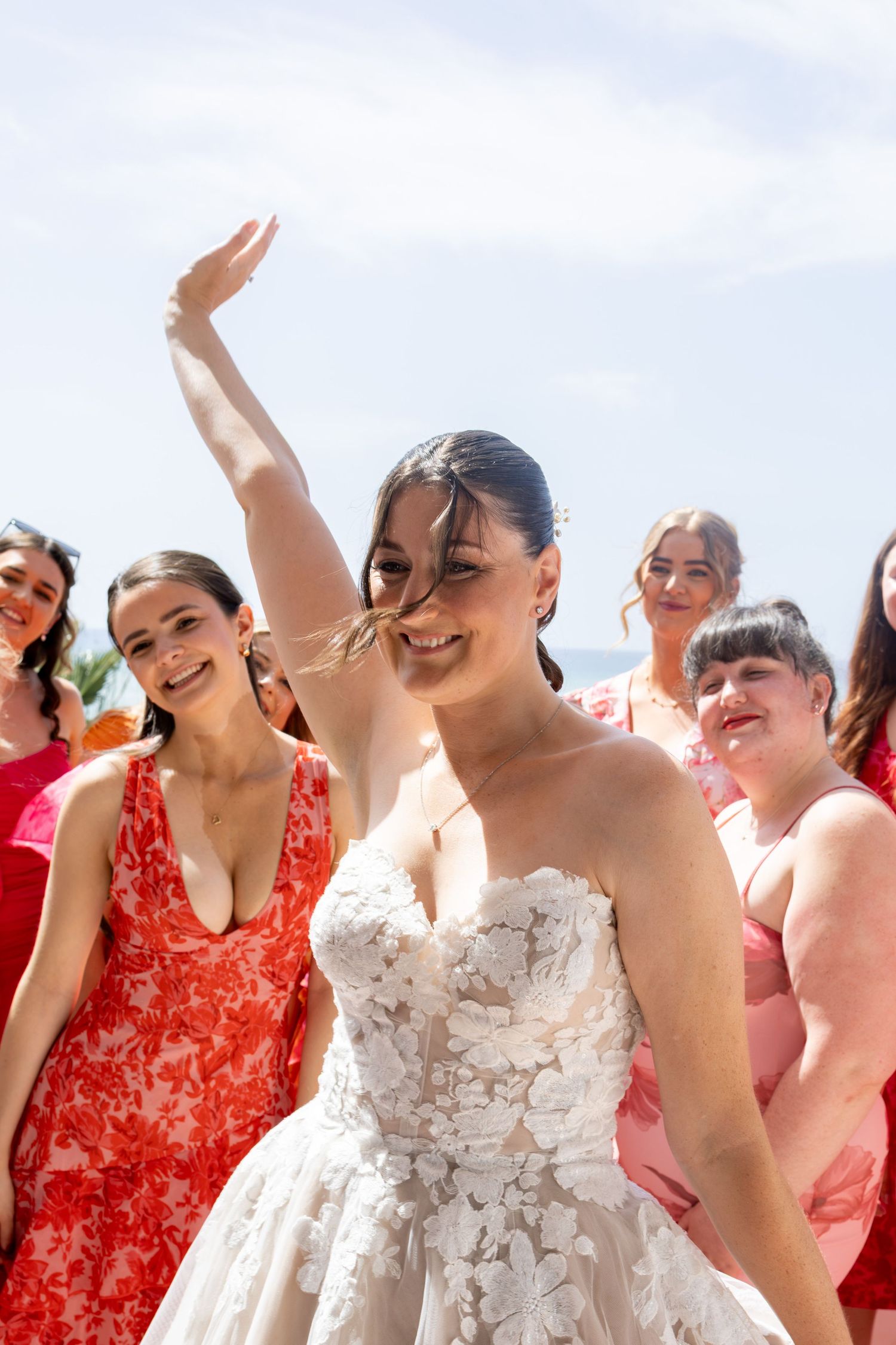 Bride in floral lace gown raises hand joyfully surrounded by guests in pink and red dresses at outdoor wedding.