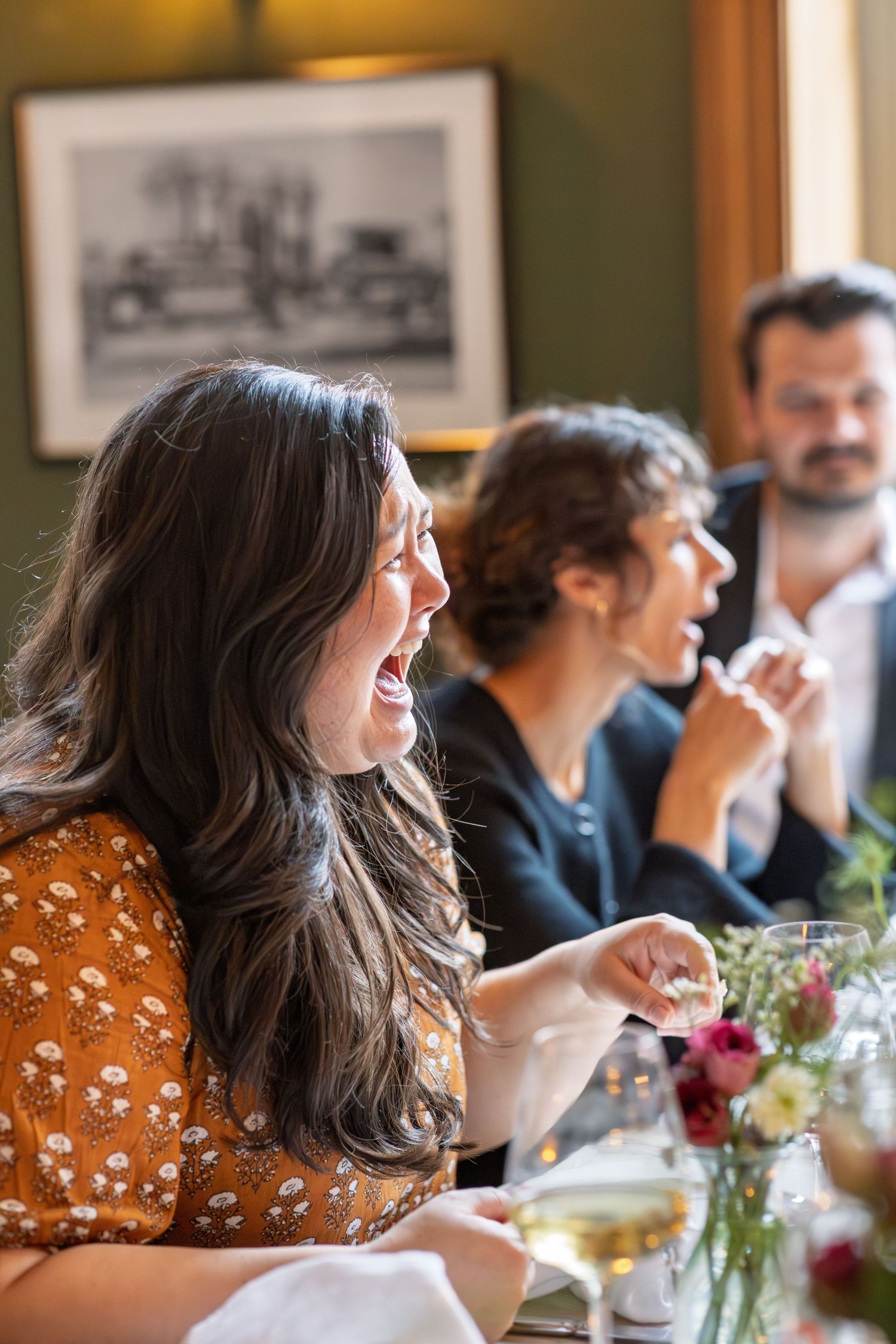 Laughing woman in floral dress enjoying wedding reception dinner with guests at decorated table.