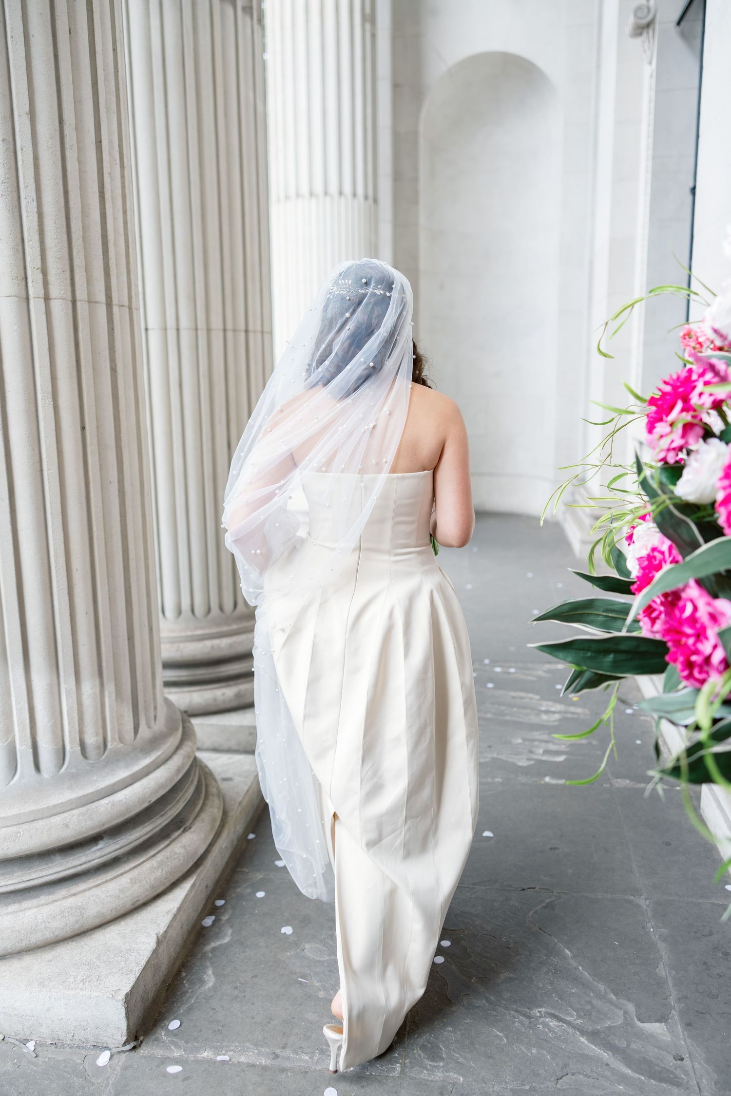 Bride in white strapless gown and veil standing beside classical stone column with pink floral arrangement nearby.